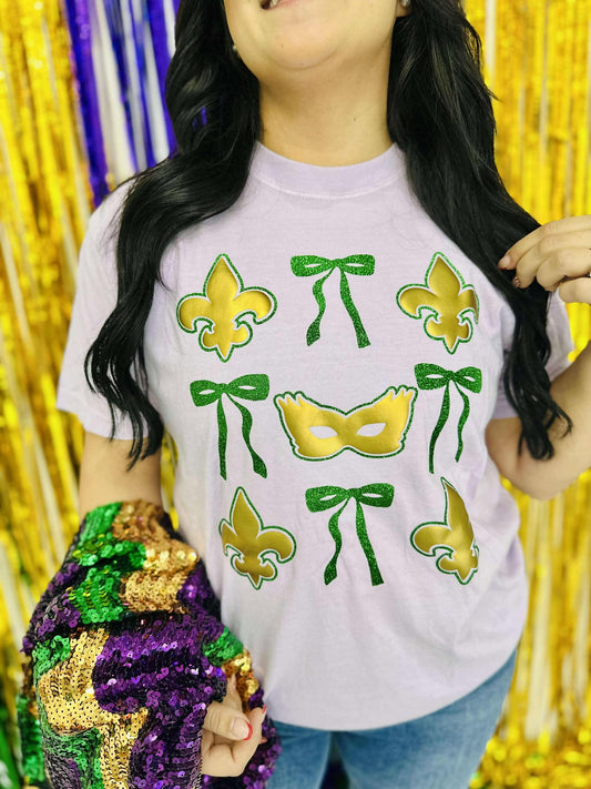 A person wearing a purple t-shirt with a green and gold bow design with masks and the text 'Mardi Gras' on it, standing in a glittery background.