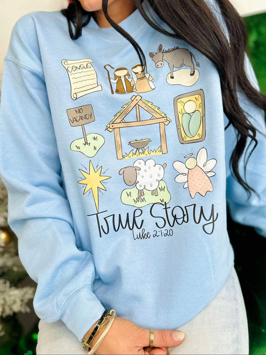 A light blue sweatshirt with a graphic design featuring a manger, sheep, and a person wearing a hat labeled 'True Story'.