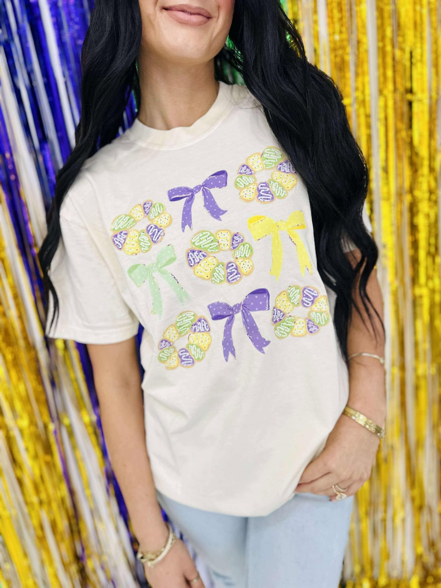 A person wearing a white graphic tee with a king cake pattern and bows, suitable for Mardi Gras celebrations.