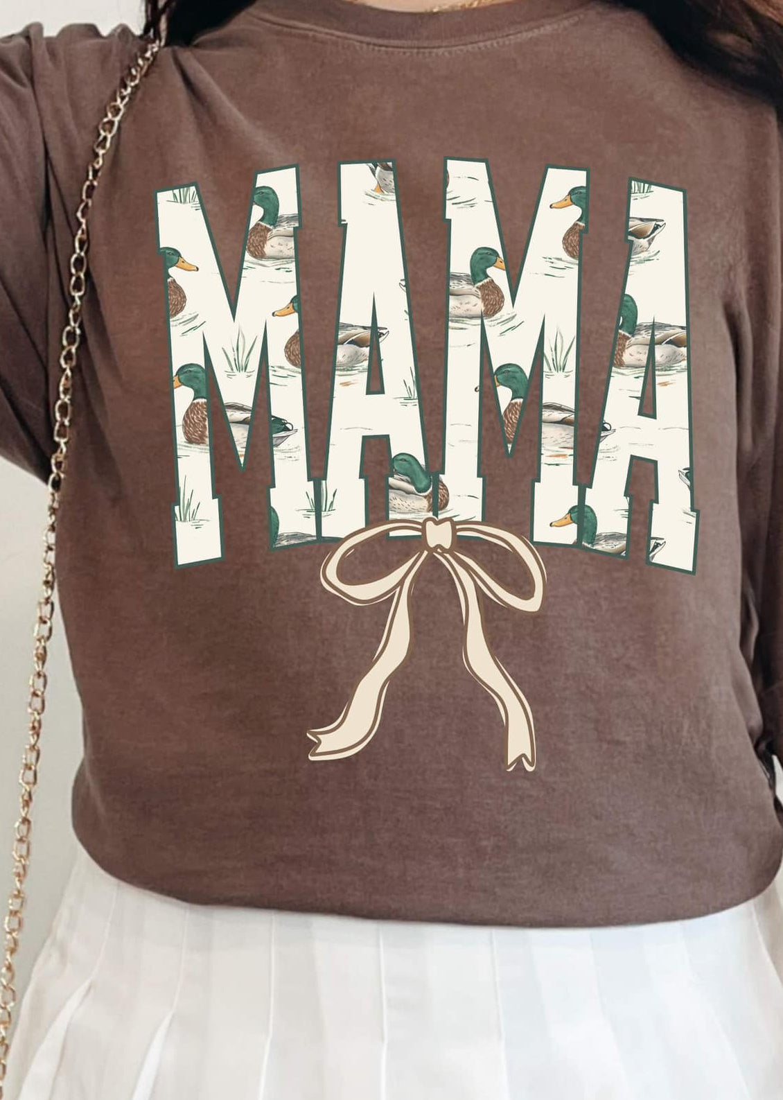 A person wearing a brown DTF tee with the word 'MAMA' and a graphic of ducks and a bow on it.