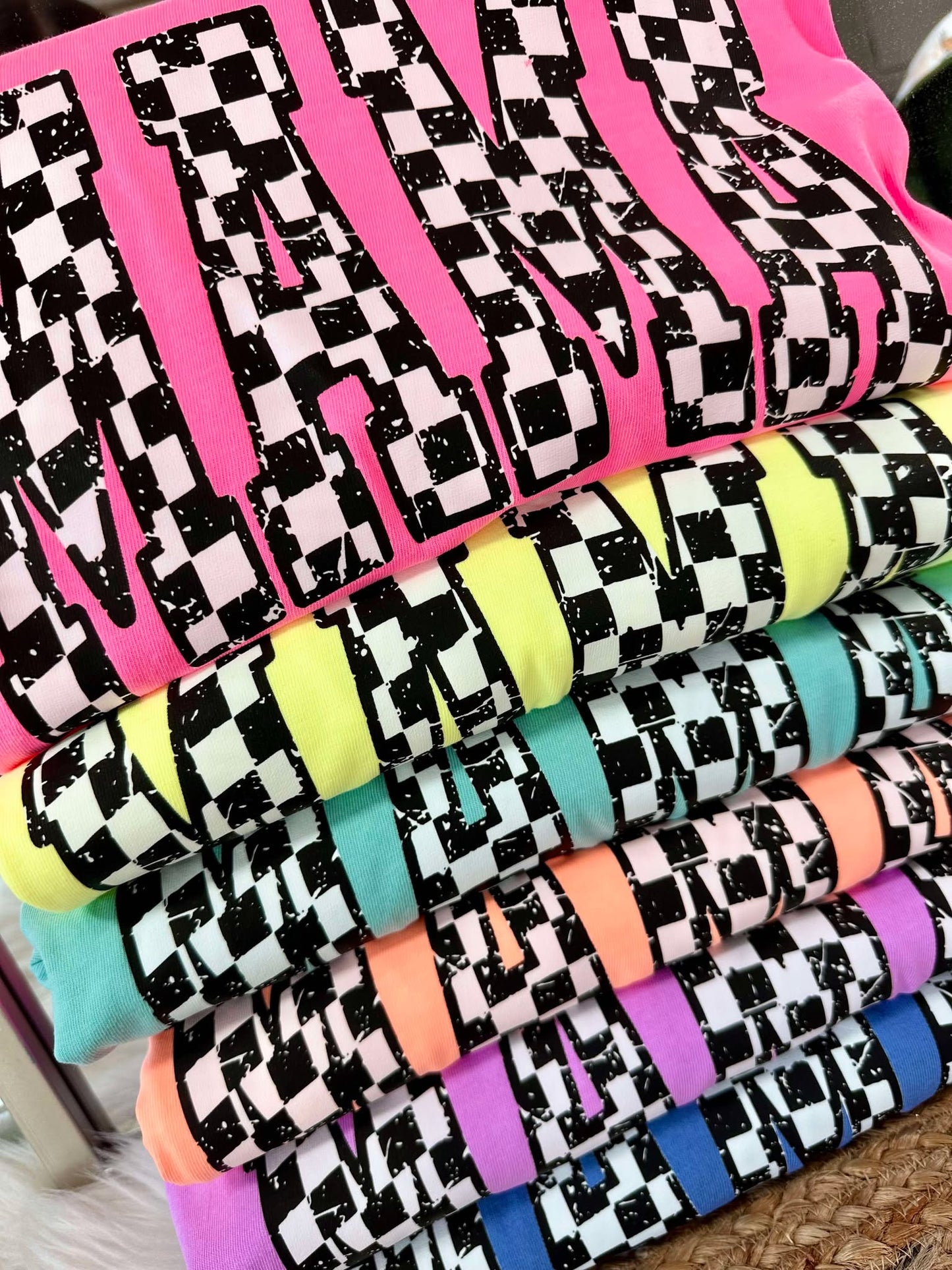 A stack of various Neon colored t-shirts with a checkered custom pattern MAMA.