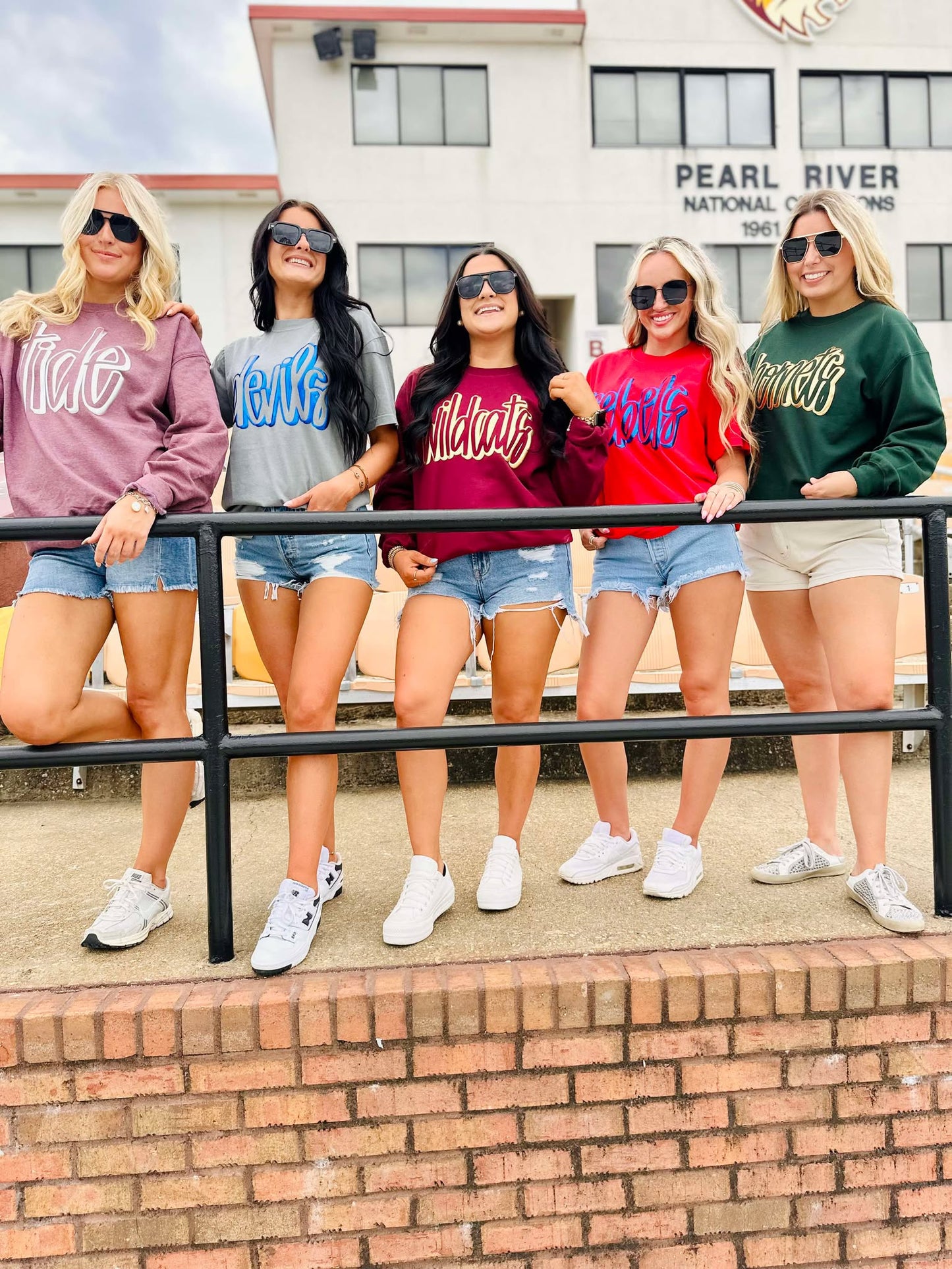 Five women posing together wearing matching sweatshirts in front of a building.