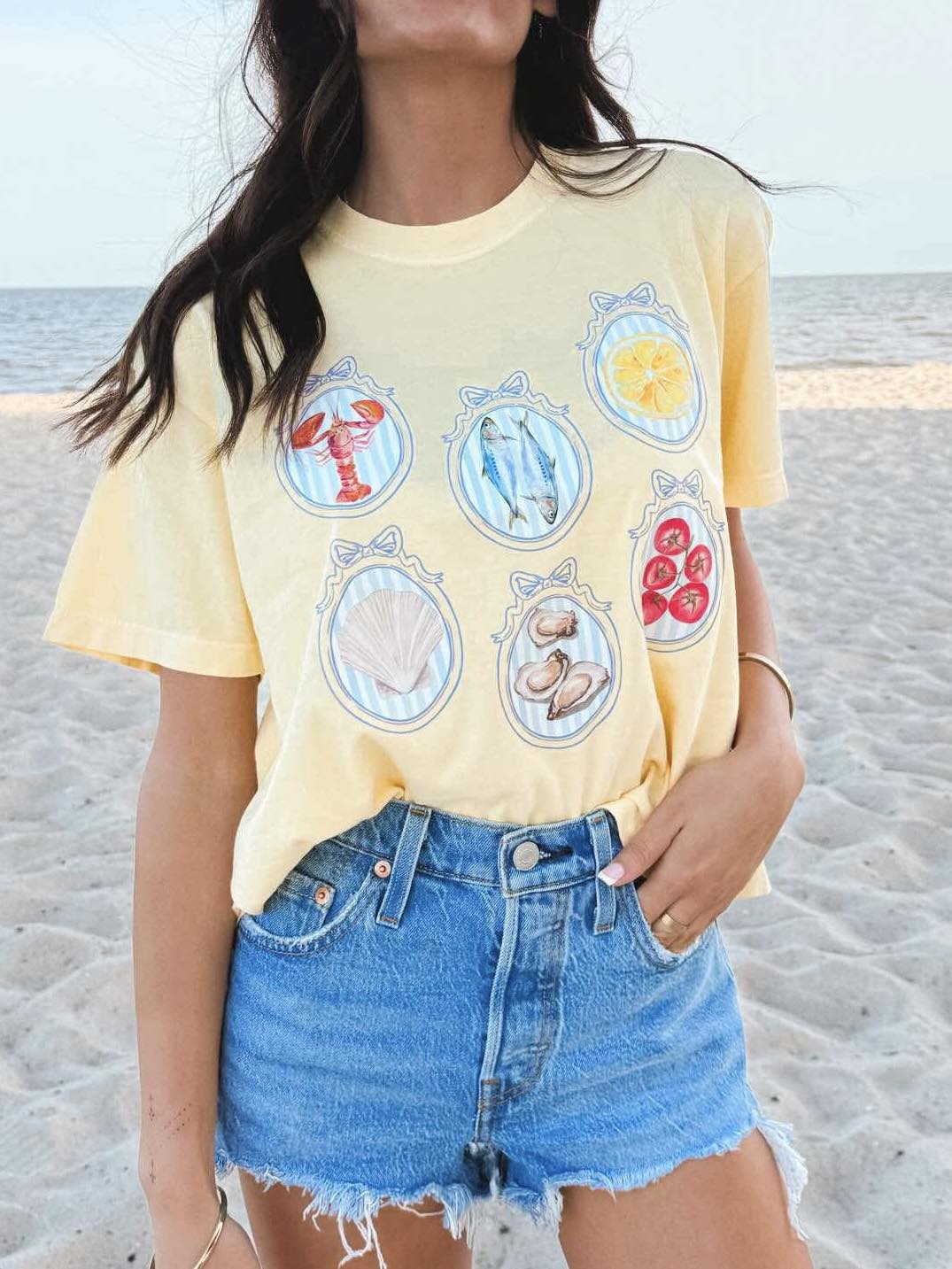 A person wearing a yellow graphic tee with seafood-themed designs and blue denim shorts, standing on a beach.