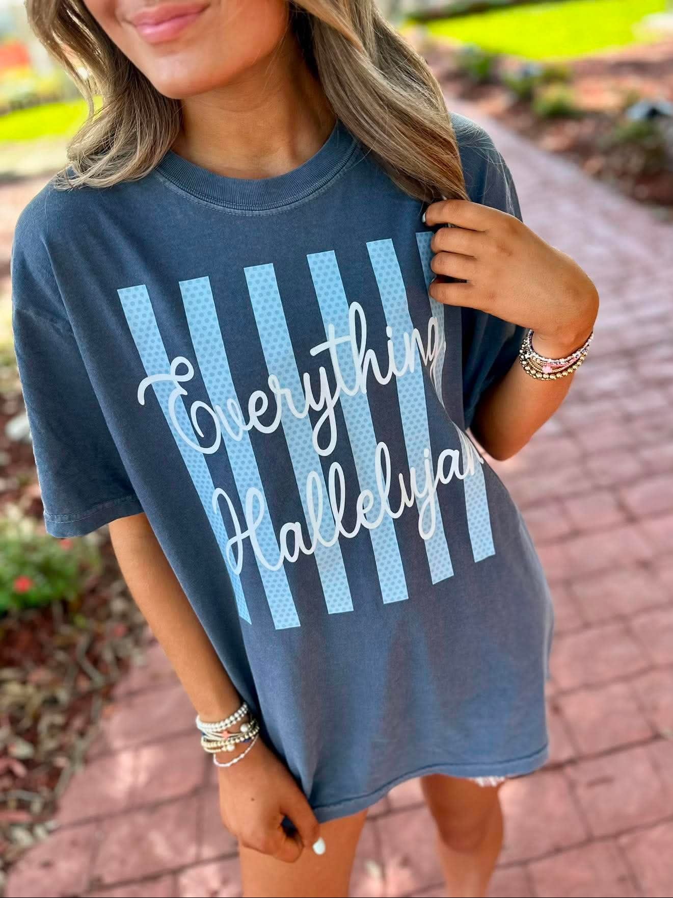 A woman wearing a slate blue graphic t-shirt with light blue vertical stripes and white cursive text reading 'Everything Hallelujah' in an outdoor setting on a brick pathway.