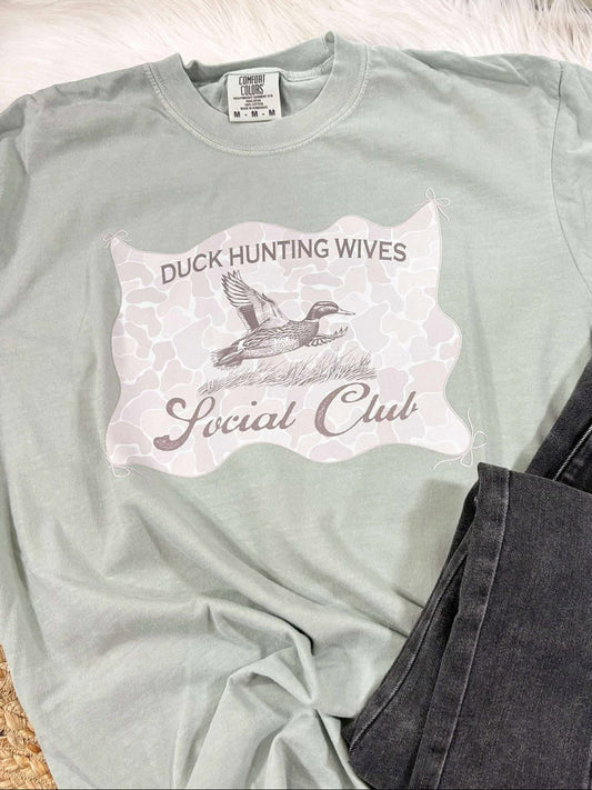 Light green t-shirt with Duck Hunting Wives Social Club graphic design on a textured surface