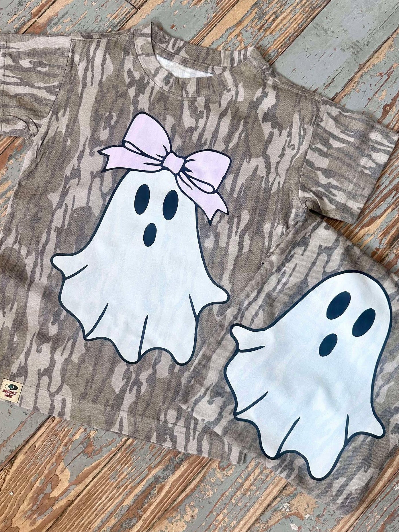 Mossy Oak Bottomland T-shirt with ghost designs and a bow on a wooden surface