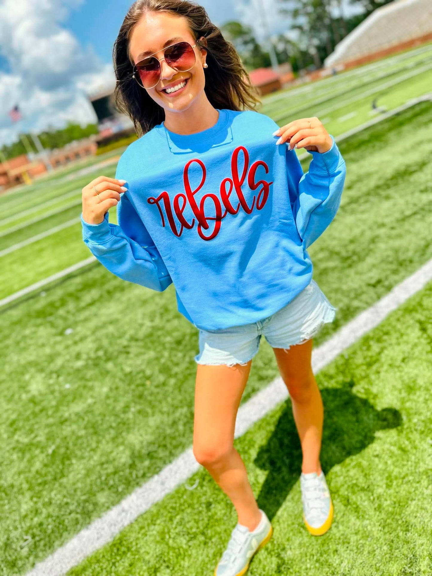 Woman wearing a blue sweatshirt with 'Rebels' on it, standing on a grassy field.