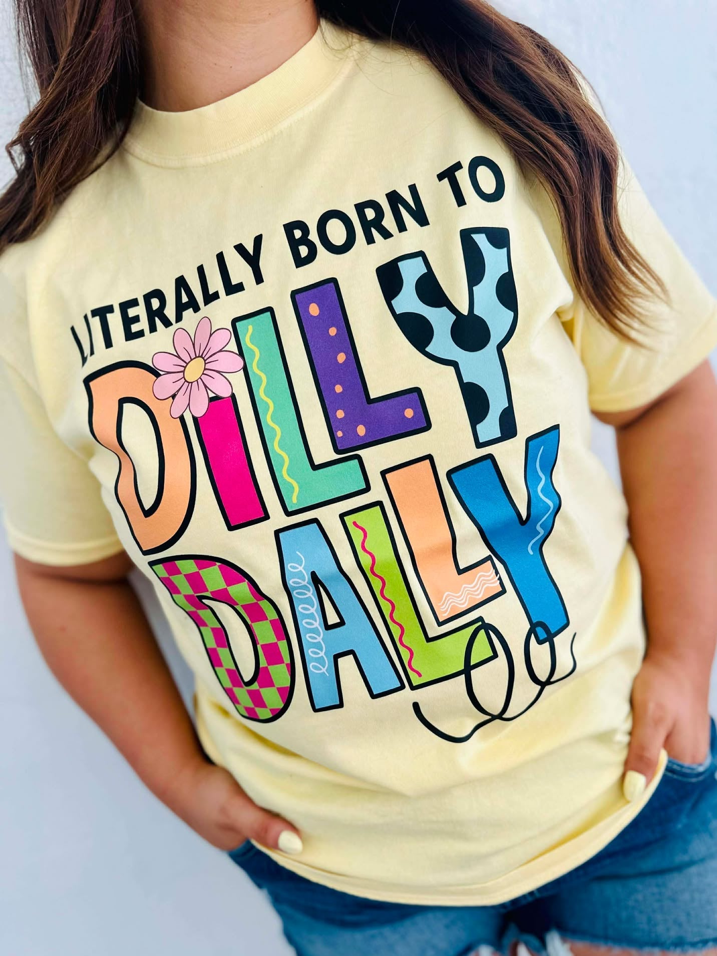 Person wearing a yellow t-shirt with colorful Born to Dilly Dally text design on a plain background