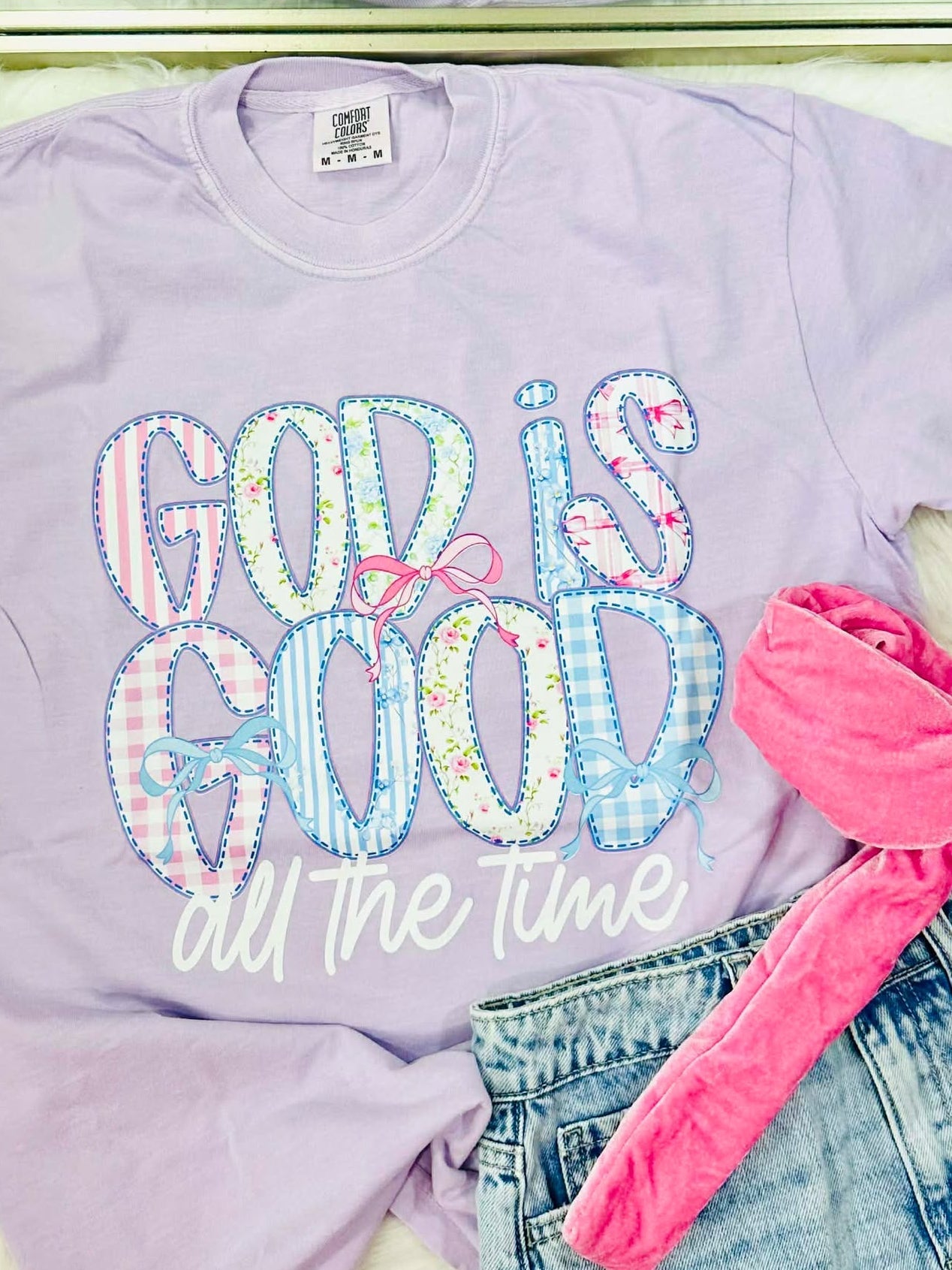 Lavender sweatshirt with 'God is good all the time' text, pink bow, and denim shorts.