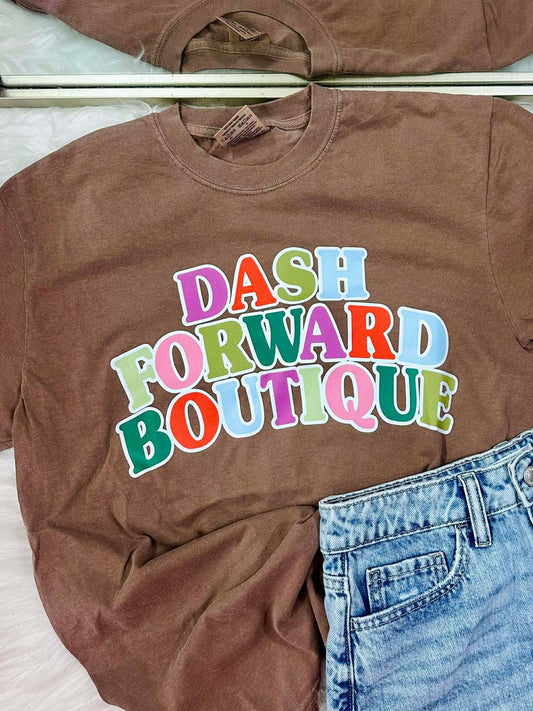 Brown t-shirt with 'Dash Forward Boutique' text and blue jeans on a light pink surface