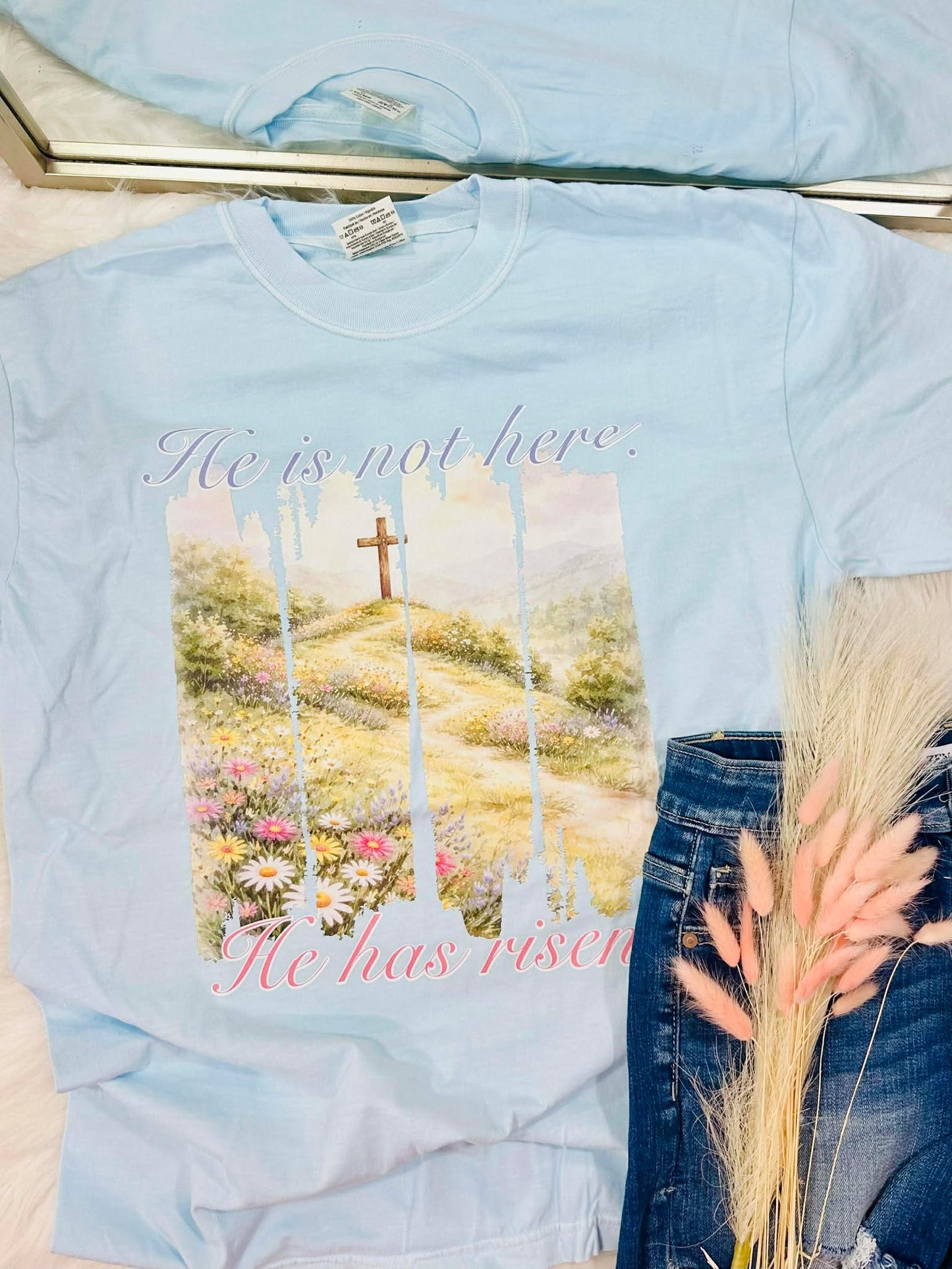 Light blue t-shirt with a printed design of a cross and flowers "He is not here. He has risen", held by a person wearing jeans.