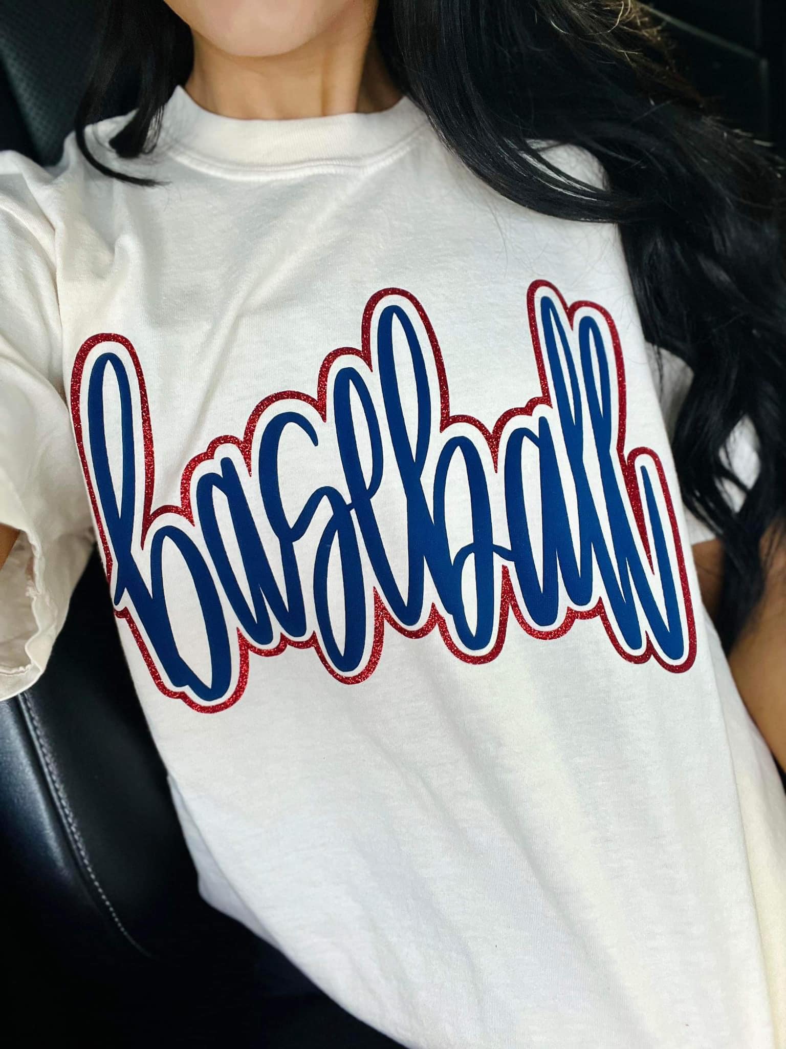 White t-shirt with 'baseball' printed in blue puff vinyl and red glitter on a person sitting in a car.