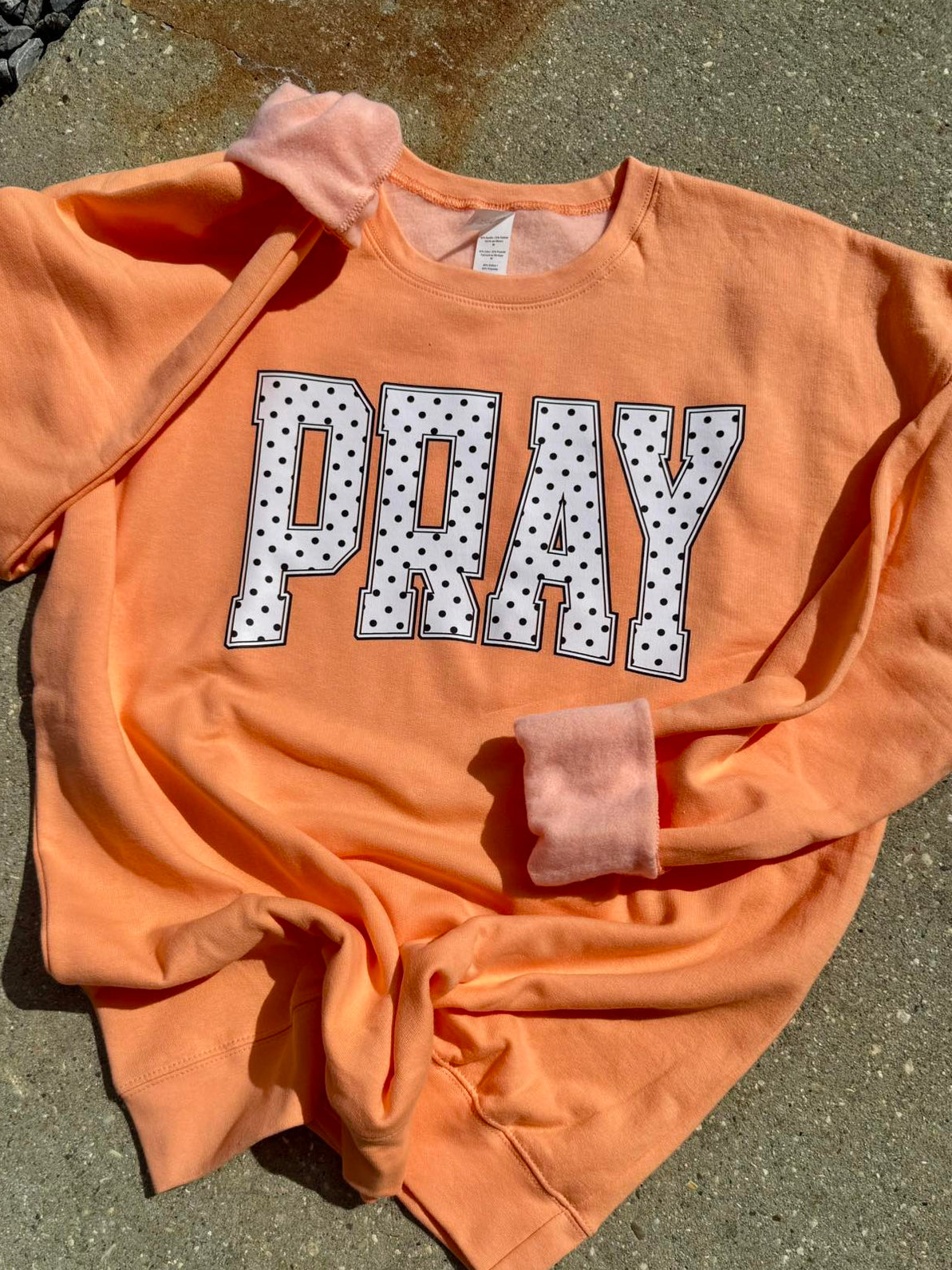 Orange sweatshirt with 'PRAY' printed on it in white letters with black polka dots, laid flat on a concrete surface.