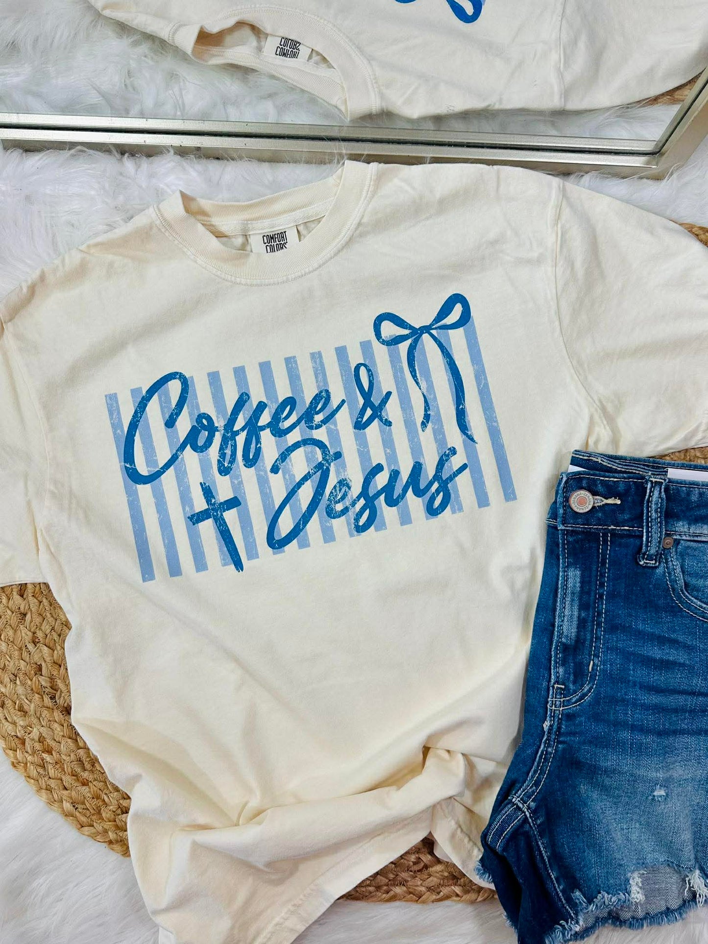White t-shirt with 'Coffee & Jesus' text and blue stripes, paired with denim shorts on a light background.