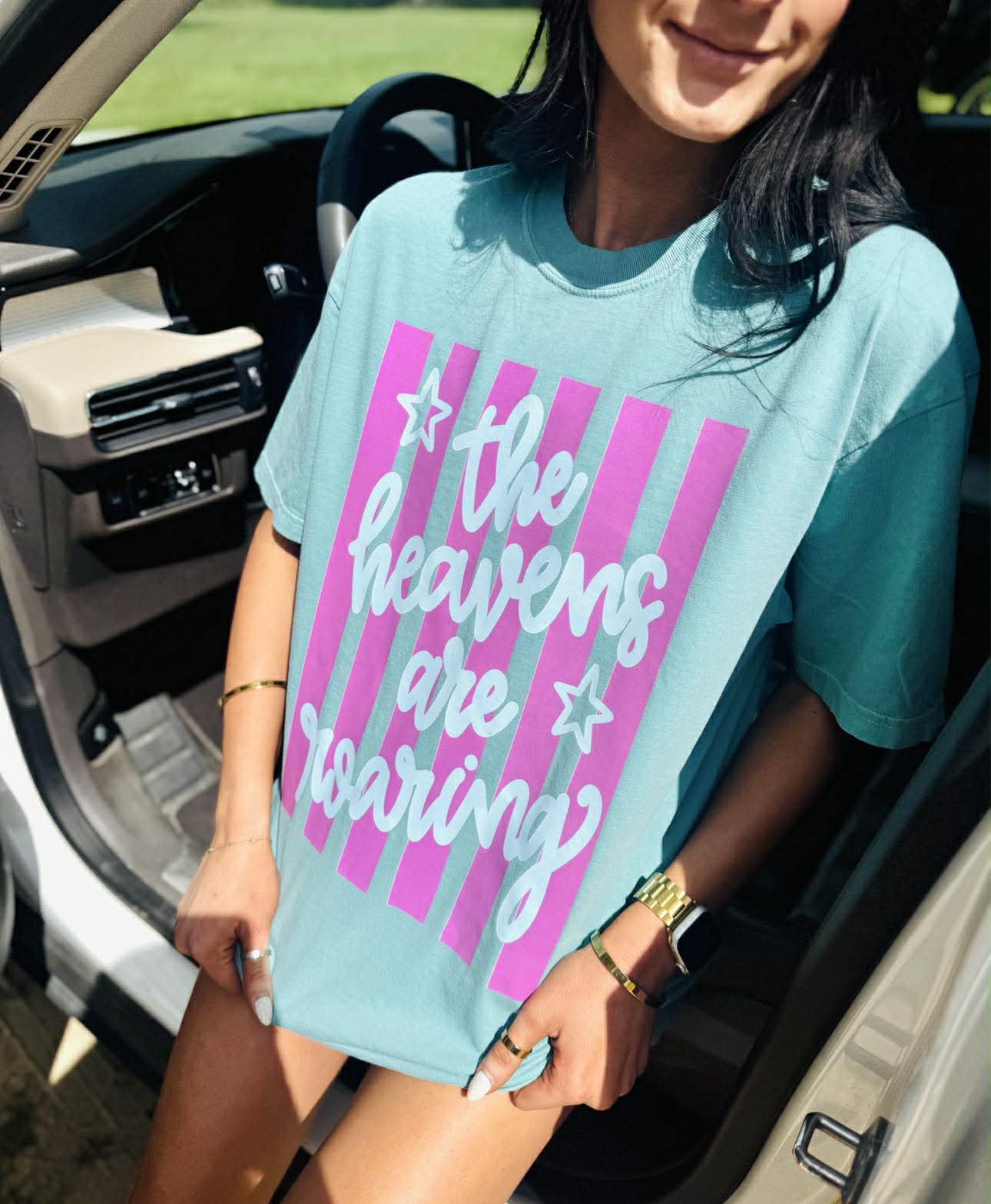 Person wearing a light blue t-shirt with pink and white text and graphics, sitting in a car.