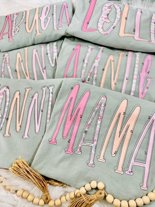 Stack of Bay Comfort Color tees with pink and white MAMA AUNTIE NONNI ABUELA NANNY LOLLI text on a light surface.