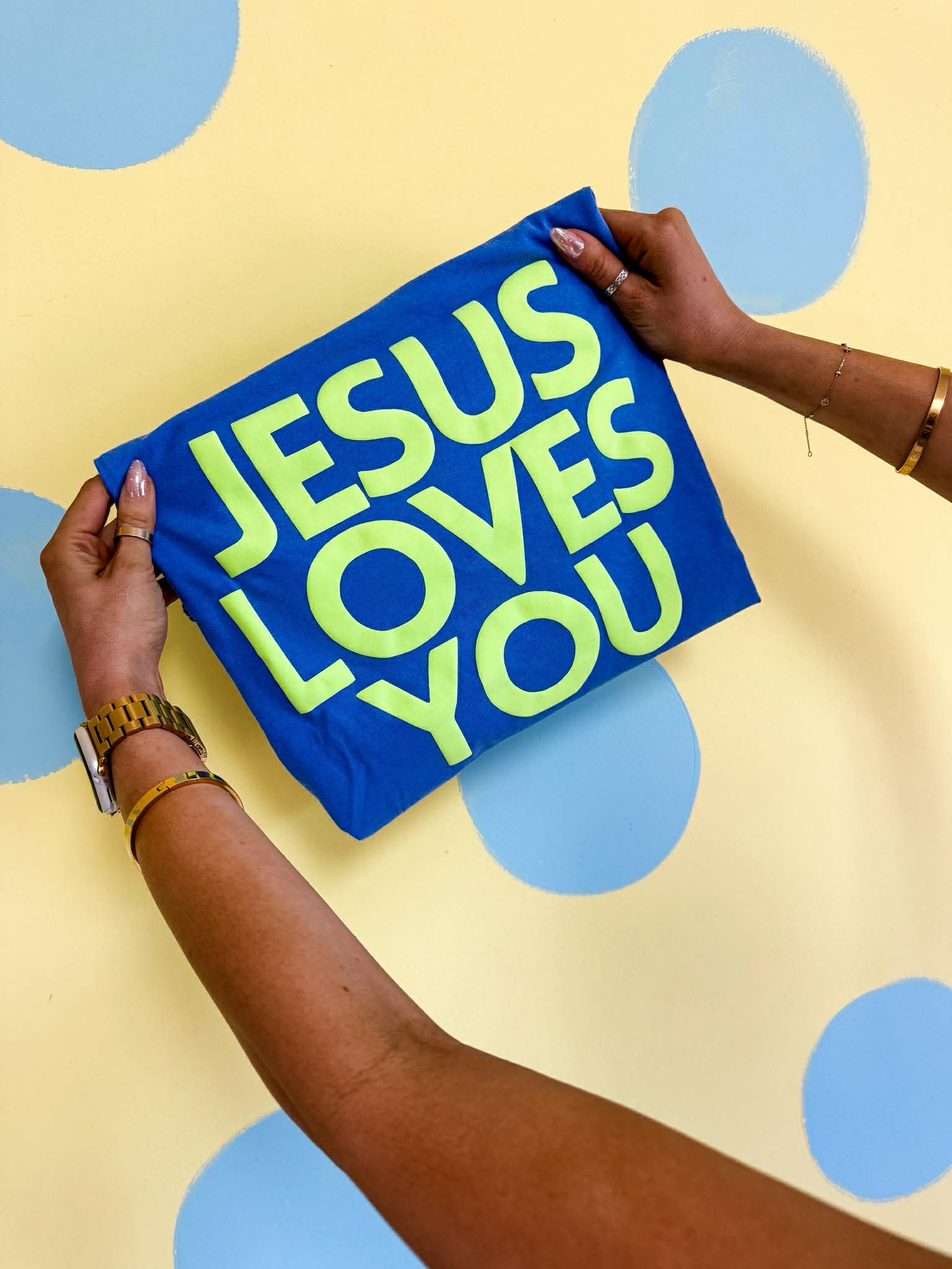 Blue pillow with 'Jesus Loves You' text held by a person against a polka dot background