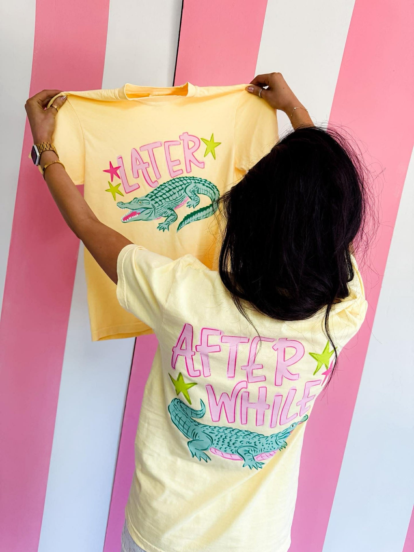 Person holding a yellow t-shirt with 'LATER' and an alligator graphic, wearing a white t-shirt with 'AFTER WHILE' and alligator graphic.