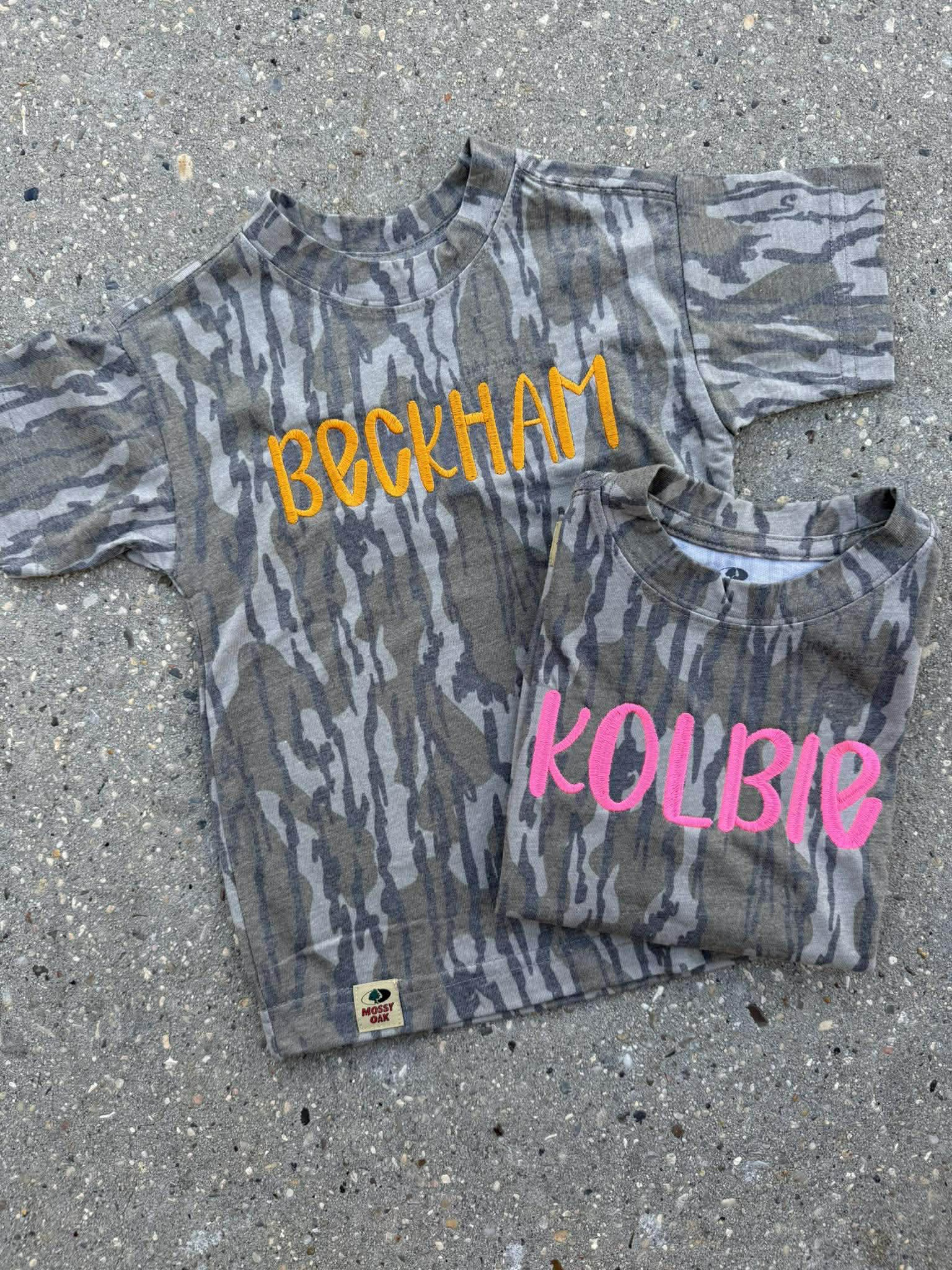 Two camouflage-patterned shirts with names 'Beckham' and 'Kolbie' on a concrete surface.