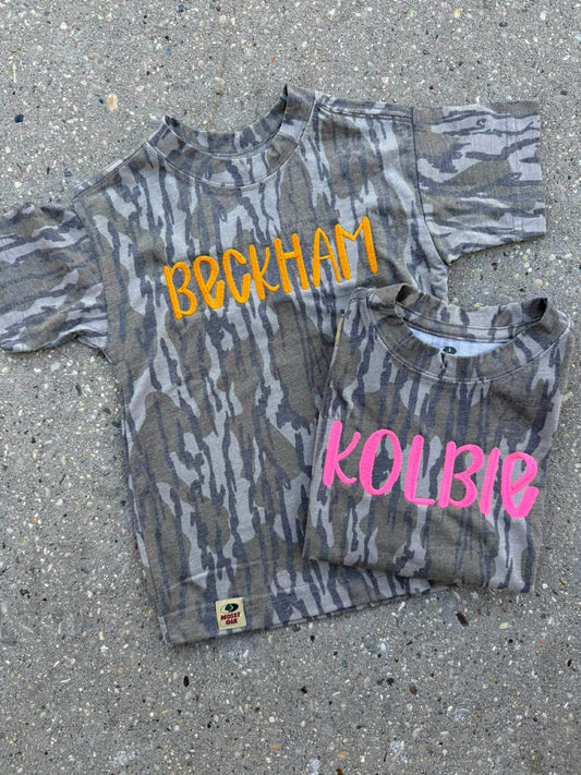 Two camouflage-patterned shirts with names 'Beckham' and 'Kolbie' on a concrete surface.