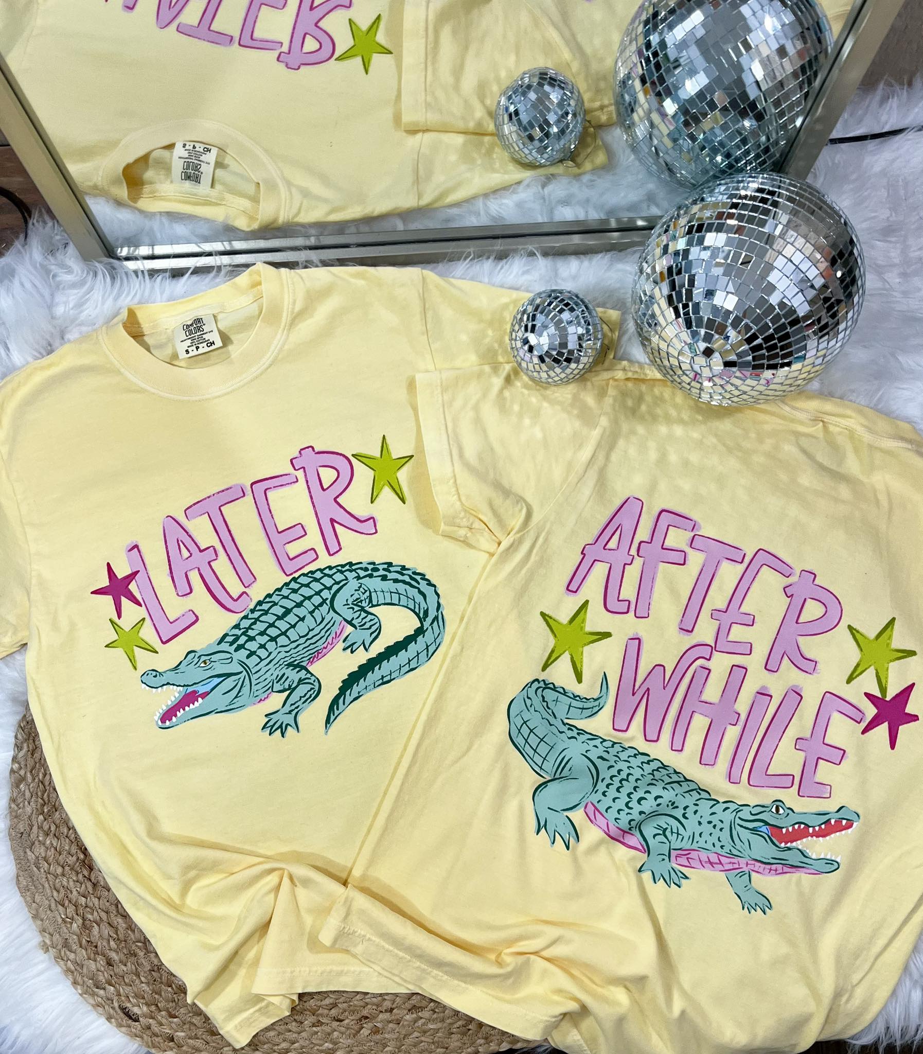 Yellow t-shirt with alligator graphic and text 'LATER' and 'AFTER WHILE' on a textured surface with disco balls.