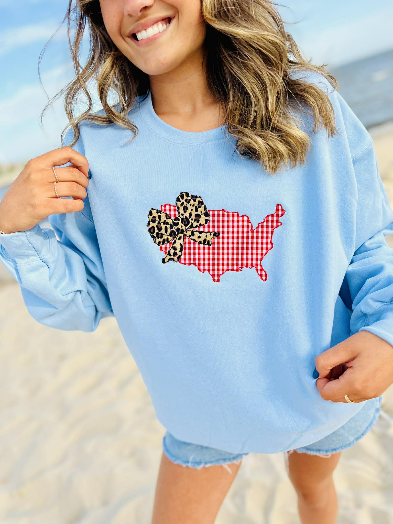 Woman wearing a light blue sweatshirt with a colorful USA map design with bow on a beach.