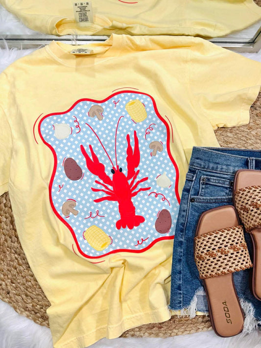 Yellow t-shirt with a red lobster design on a woven surface with jeans and sandals.