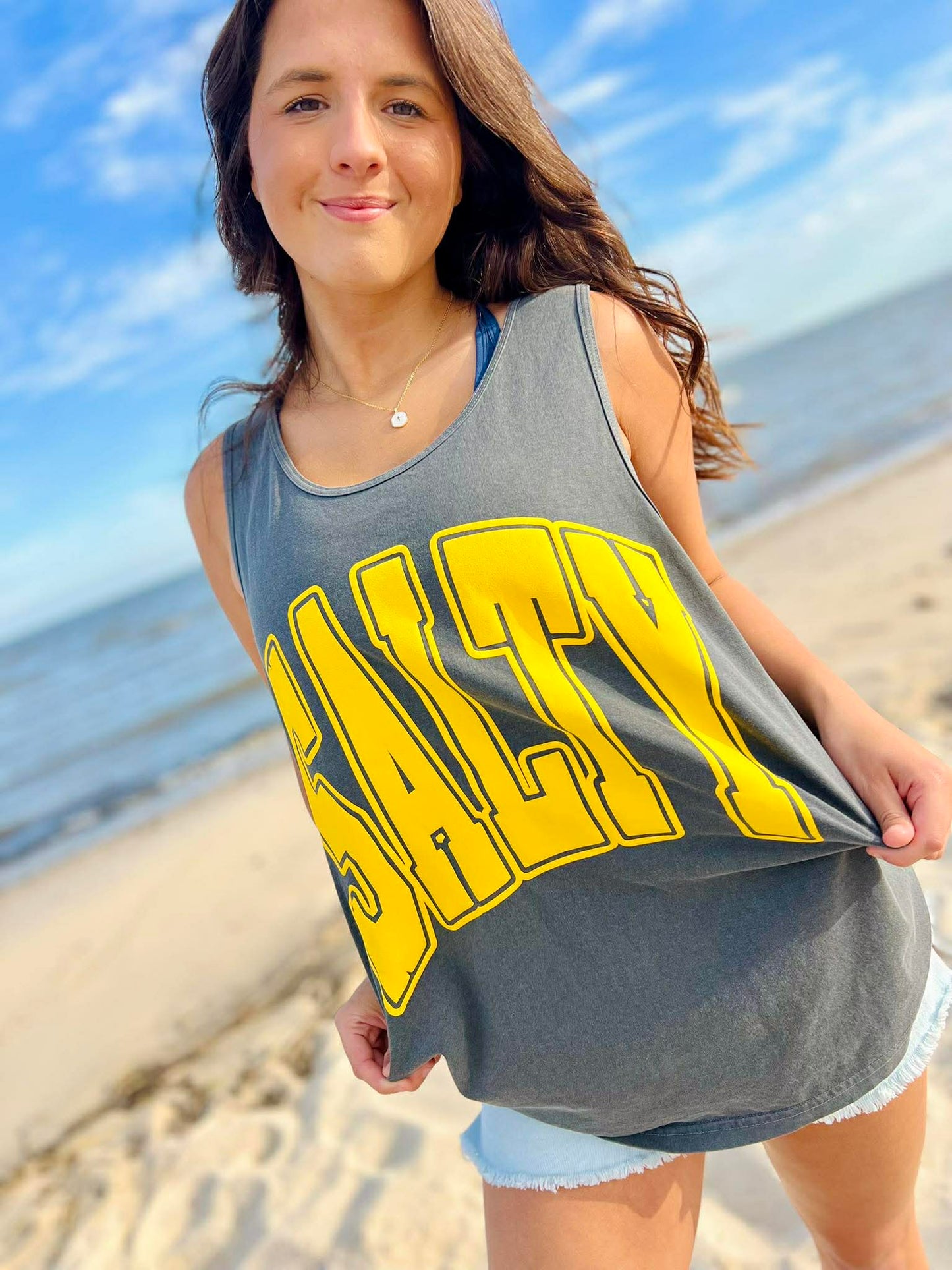 Woman wearing a gray tank top with 'Salty' printed in yellow on a beach.