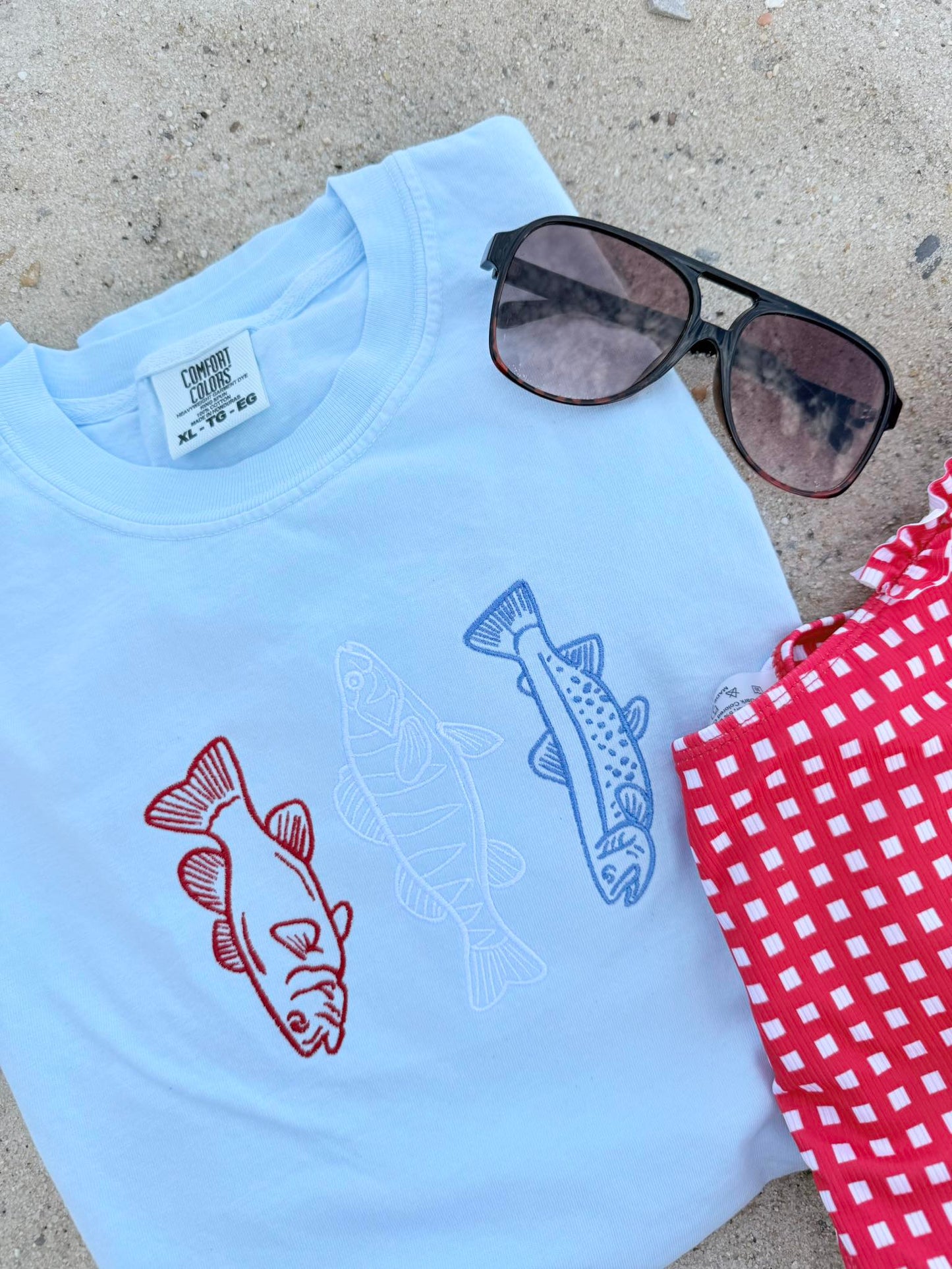 Light blue t-shirt with fish designs, sunglasses, and a red checkered cloth on a concrete surface.