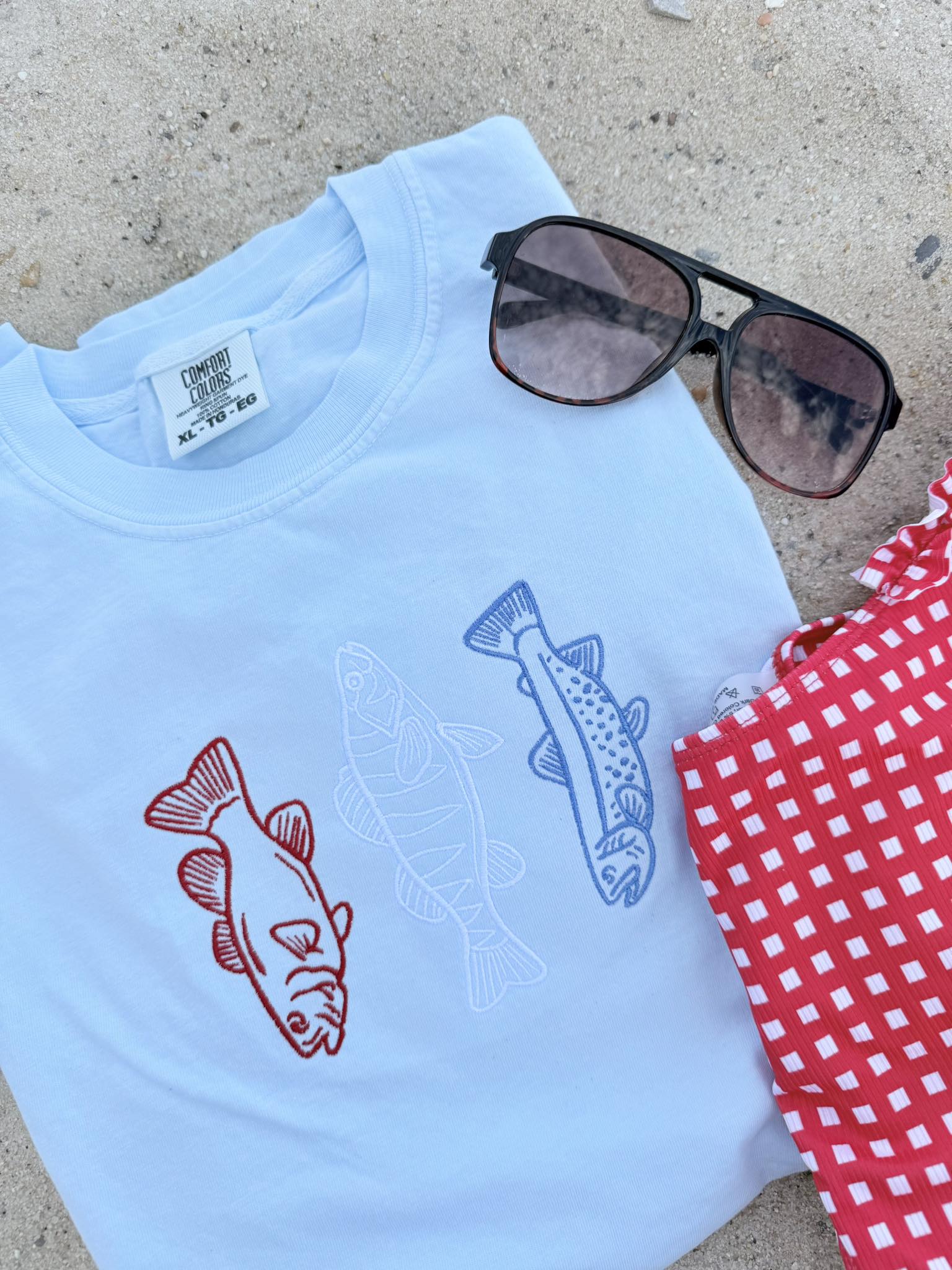 Light blue t-shirt with fish designs, sunglasses, and a red checkered cloth on a concrete surface.