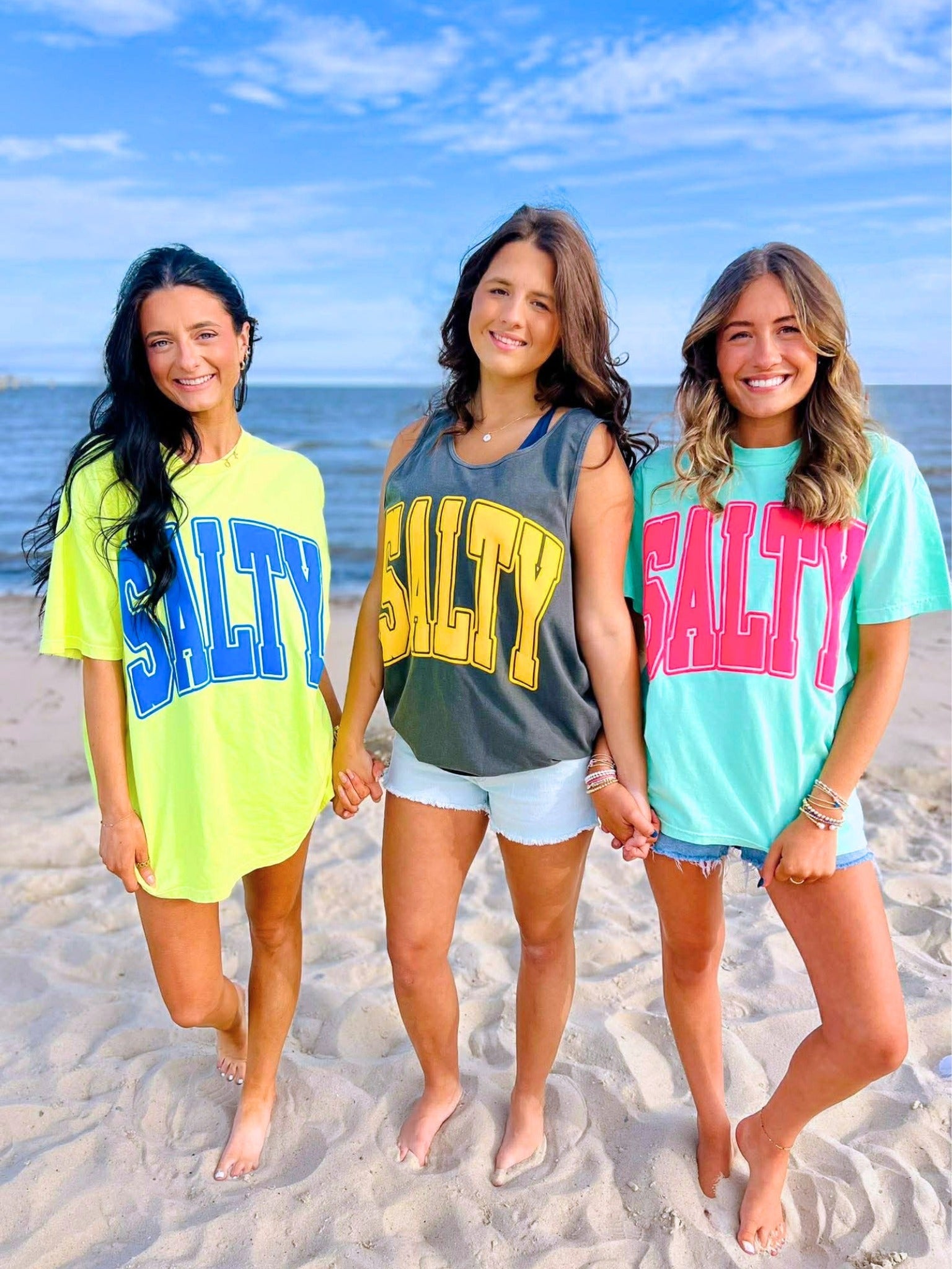 Three women on a beach wearing 'SALTY' t-shirts in different colors.