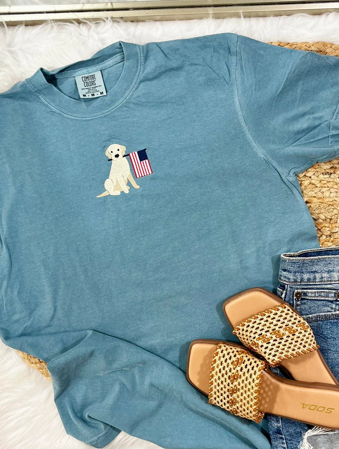 Blue t-shirt with a dog and American flag design, paired with sandals and jeans on a white surface.