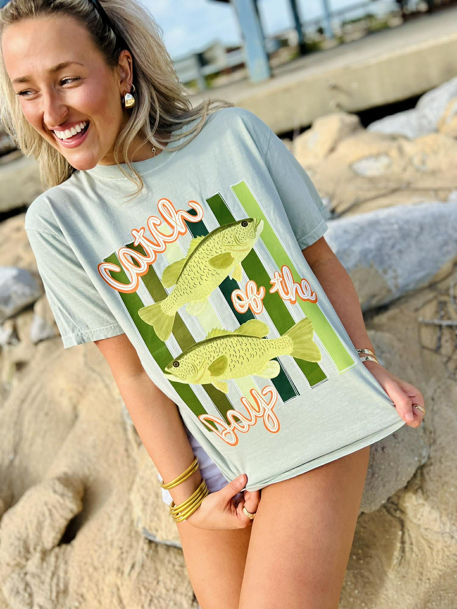 Woman wearing a light blue t-shirt with fish graphics and text on a rocky beach.