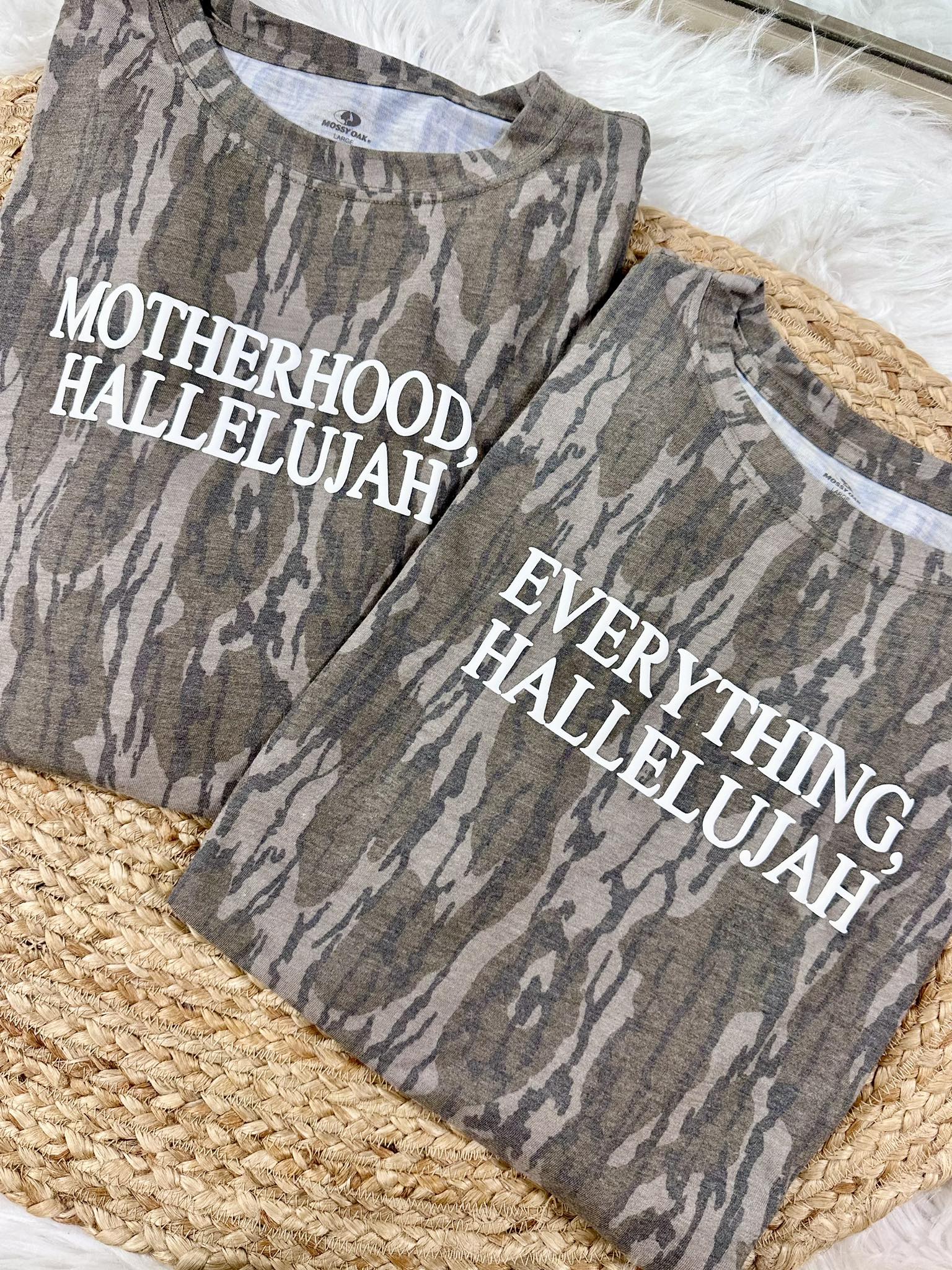 Two camouflage-patterned shirts with Motherhood, Hallelujah and Everything, Hallelujah puff text on a Bottomland surface
