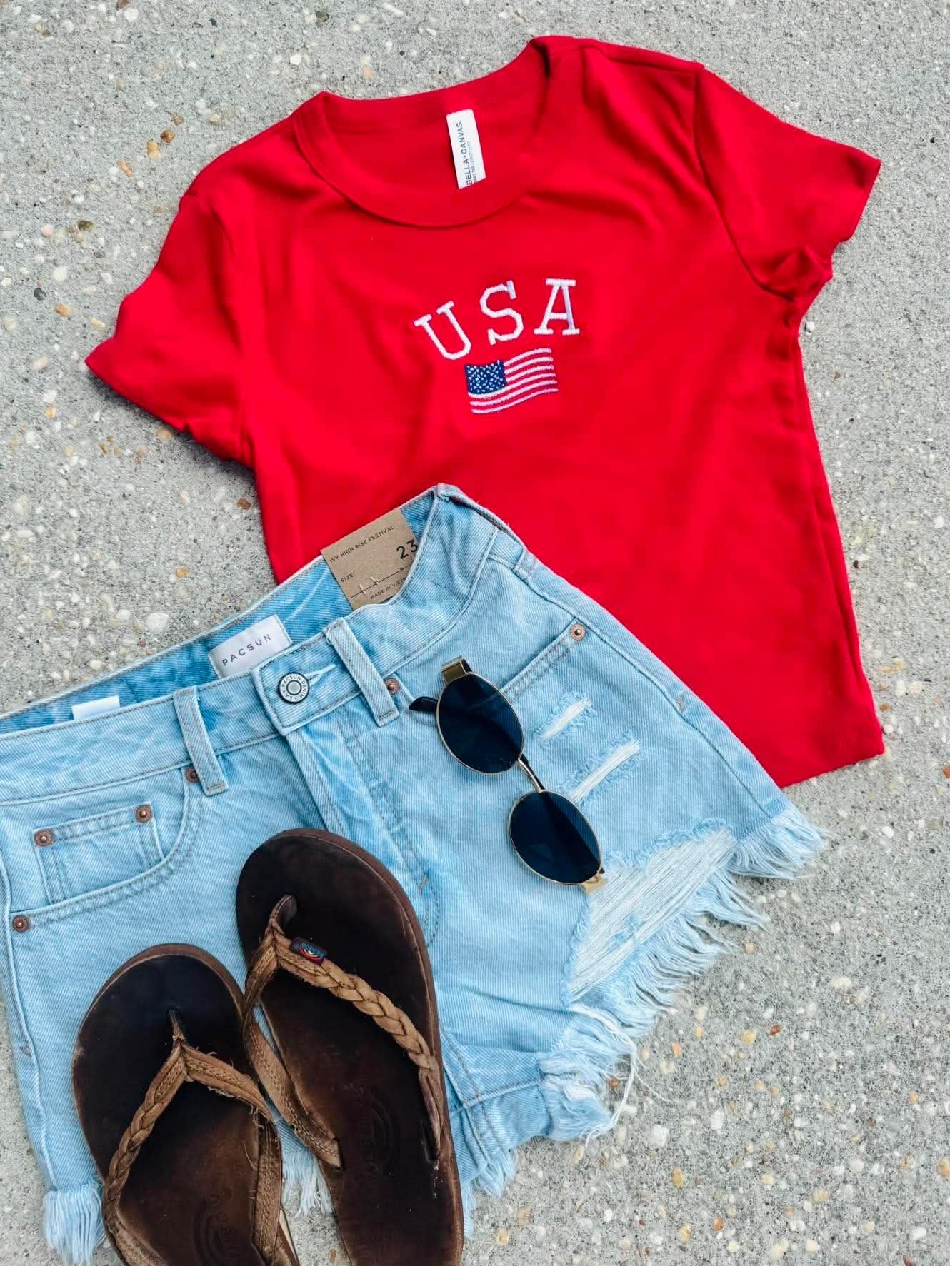 Red 'USA' t-shirt with denim shorts and sandals on a concrete surface