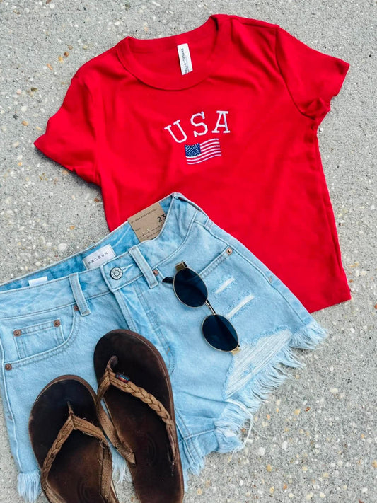 Red 'USA' t-shirt with denim shorts and sandals on a concrete surface