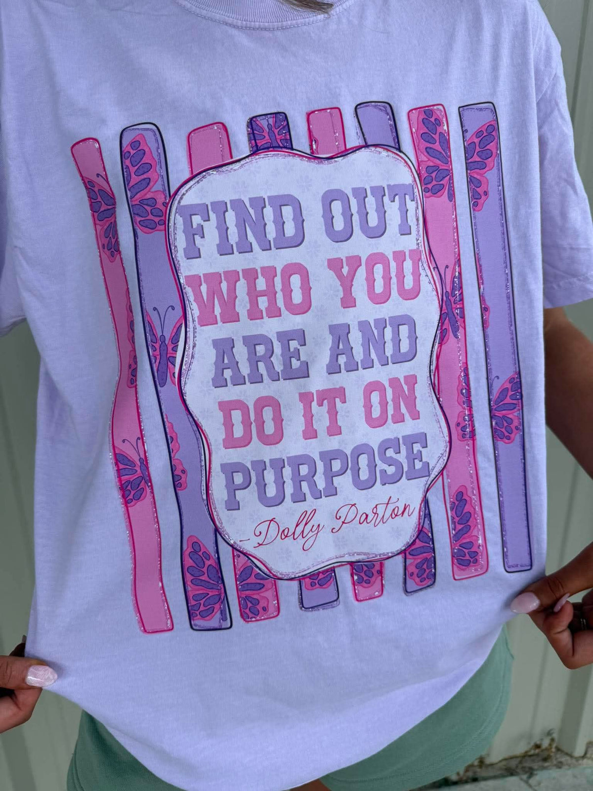 A person wearing a white graphic T-shirt with a pink and purple fence design and the text 'FIND OUT WHO YOU ARE AND DO IT ON PURPOSE' with 'Dolly Parton' written below, photographed outdoors against a light green wall.