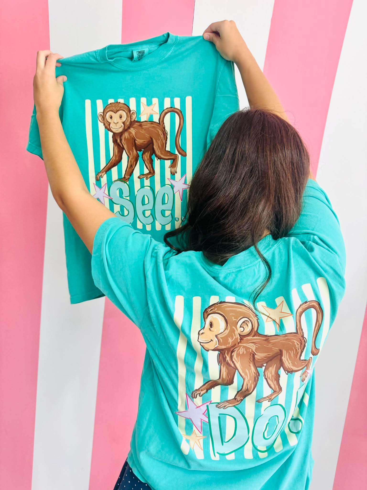 Person holding a turquoise t-shirt with monkey see monkey do striped graphics against a pink and white striped background