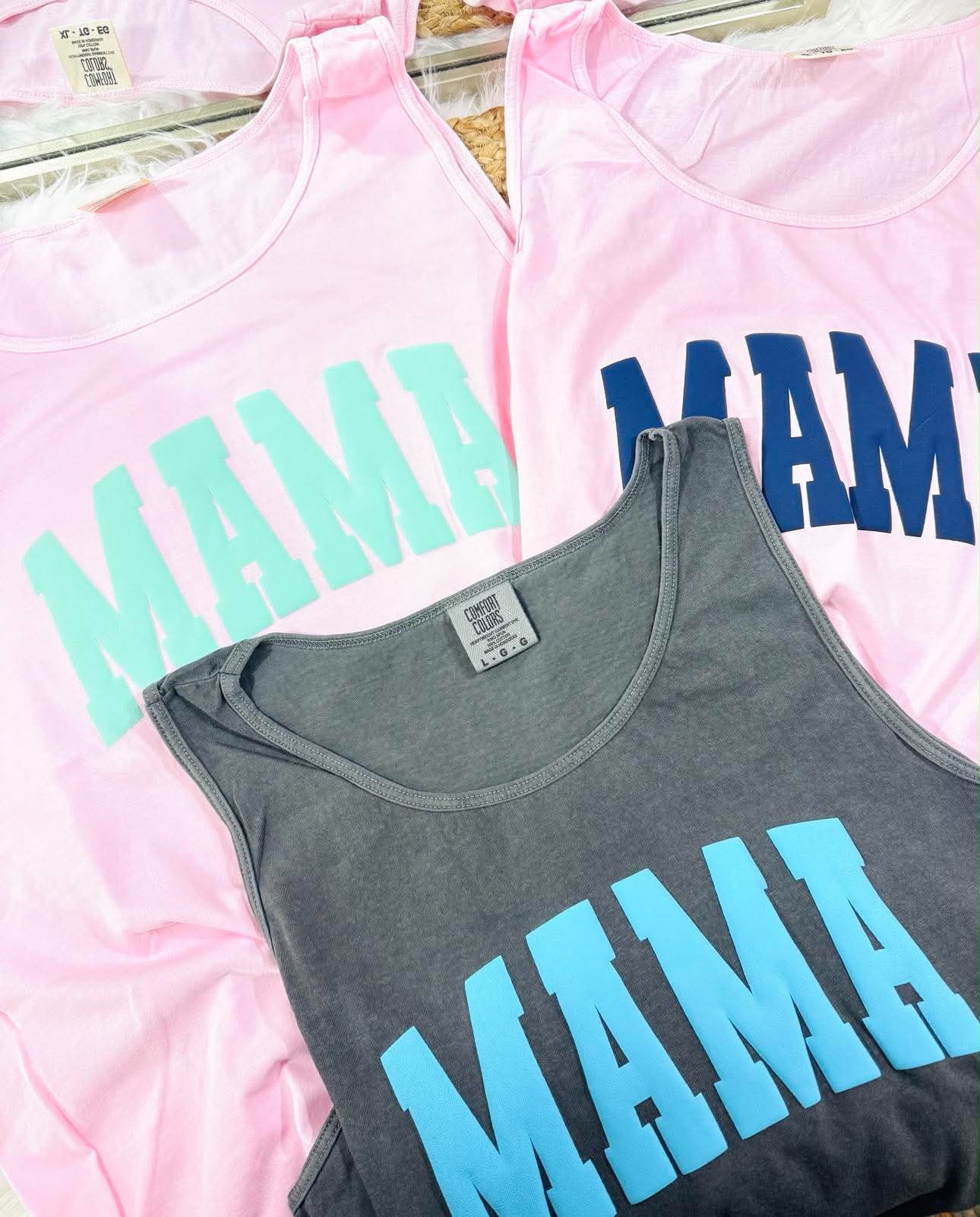 Three tank tops with 'MAMA' text in different colors on a pink background.