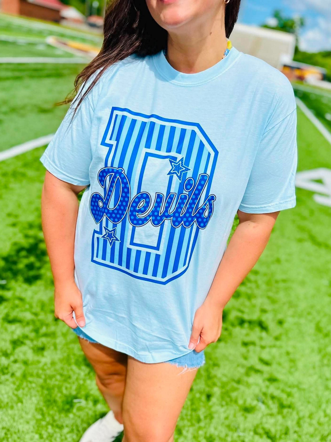 Person wearing a light blue t-shirt with 'Devils' printed on it, standing on a grassy field.