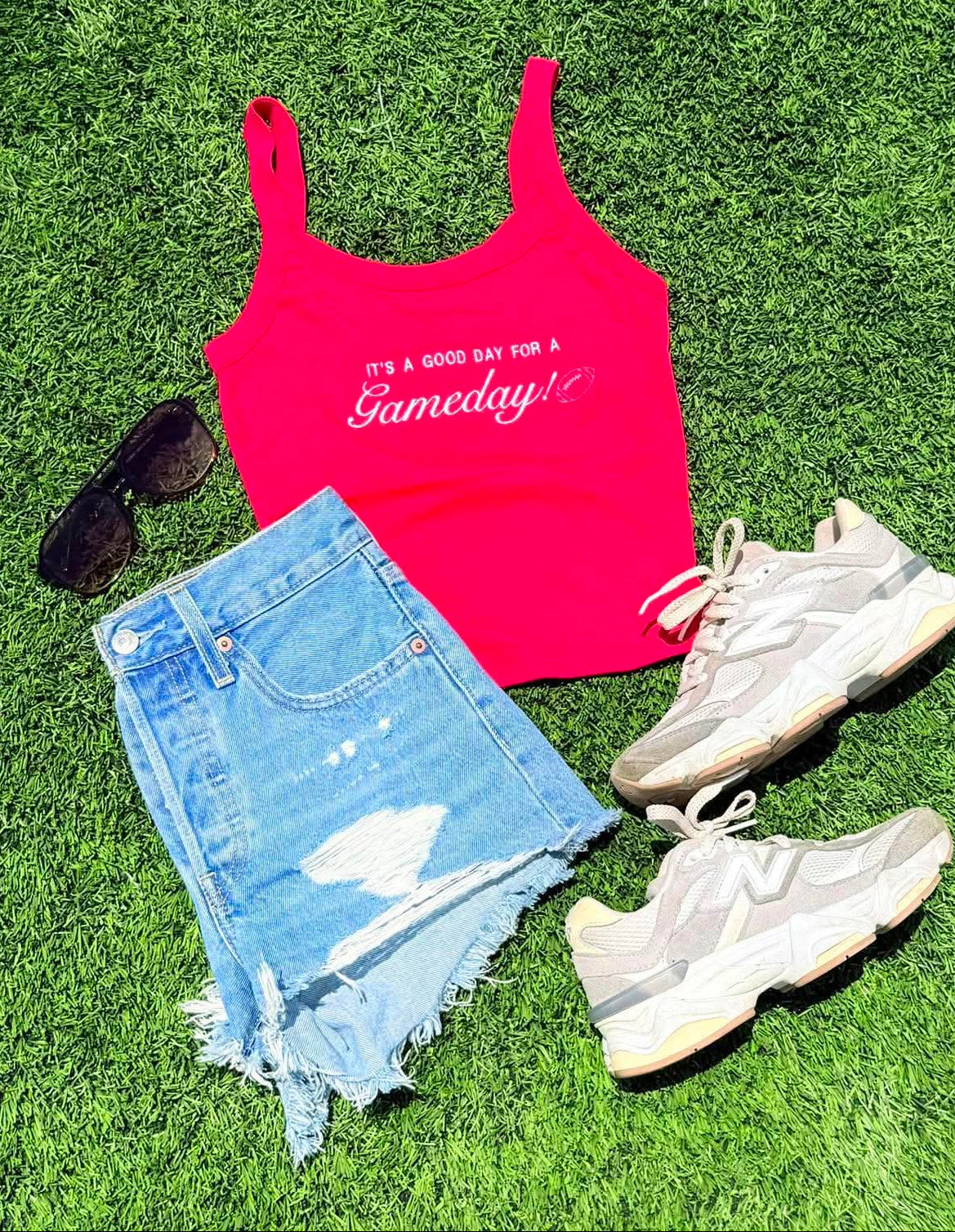 Pink tank top with text, denim shorts, sneakers, and sunglasses on grass