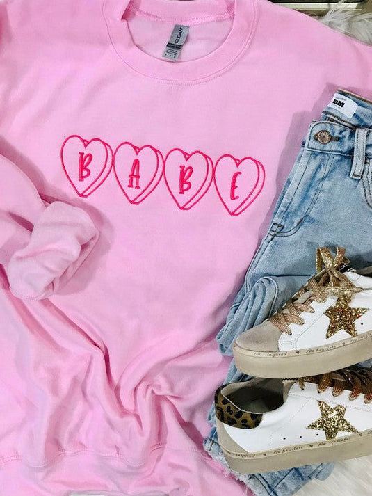 A pink embroidered sweatshirt with the word 'BABE' in heart shape on the front, displayed flat with a pair of blue jeans and white sneakers on top.