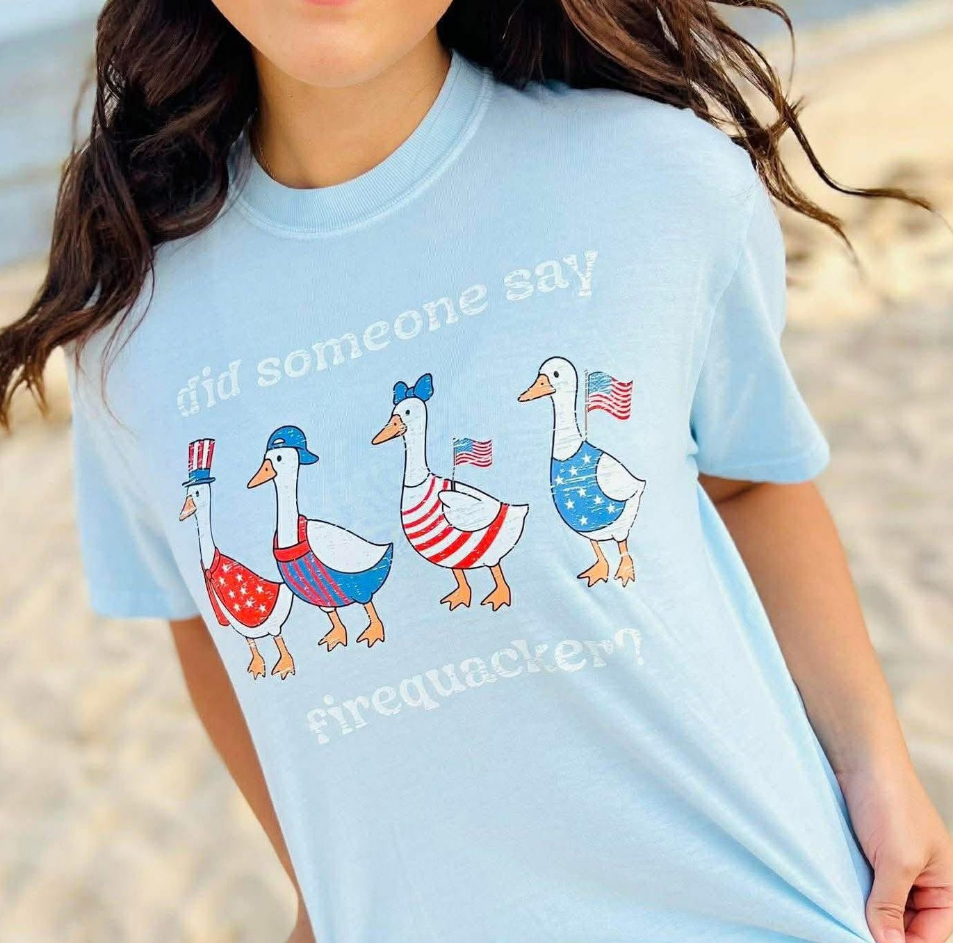 Light blue t-shirt with patriotic duck graphics worn by a person on a beach.