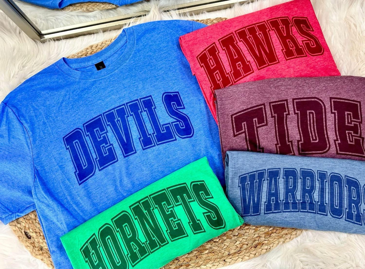 Collection of t-shirts with 'DEVILS', 'HAWKS', 'TIDE', and 'WARRIORS' text on a textured surface.