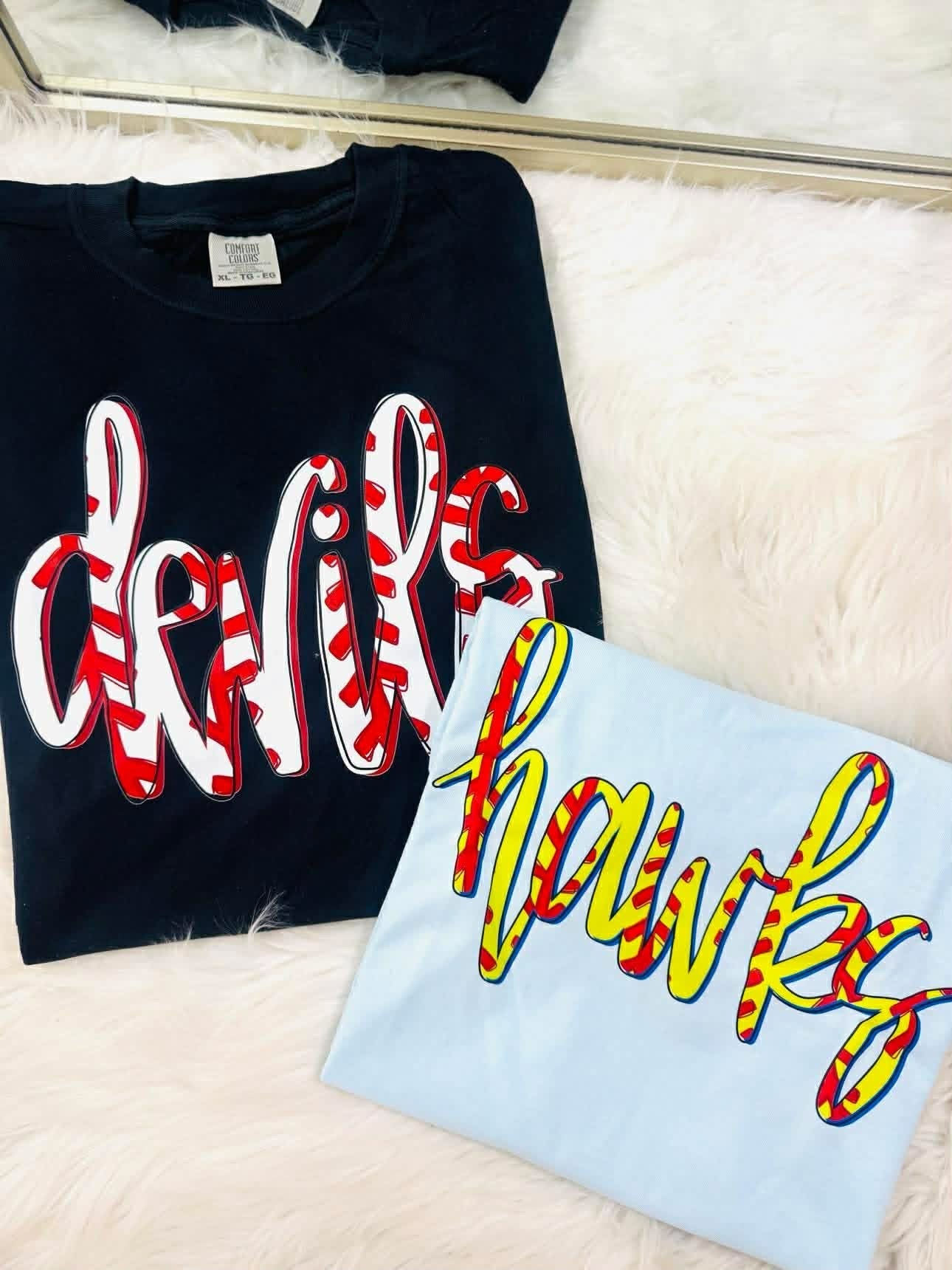 Black shirt with red and white text and light blue shirt with yellow and red text on a white surface.