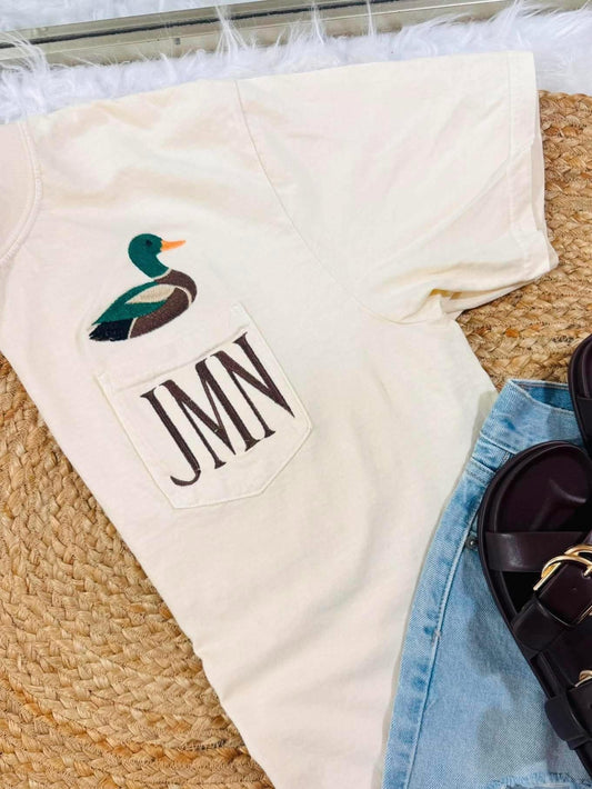 An off-white short-sleeve t-shirt with a front pocket featuring a duck graphic and the monogram 'JMN', styled in a flat lay with light-wash denim shorts and dark brown slide sandals on a woven rug.