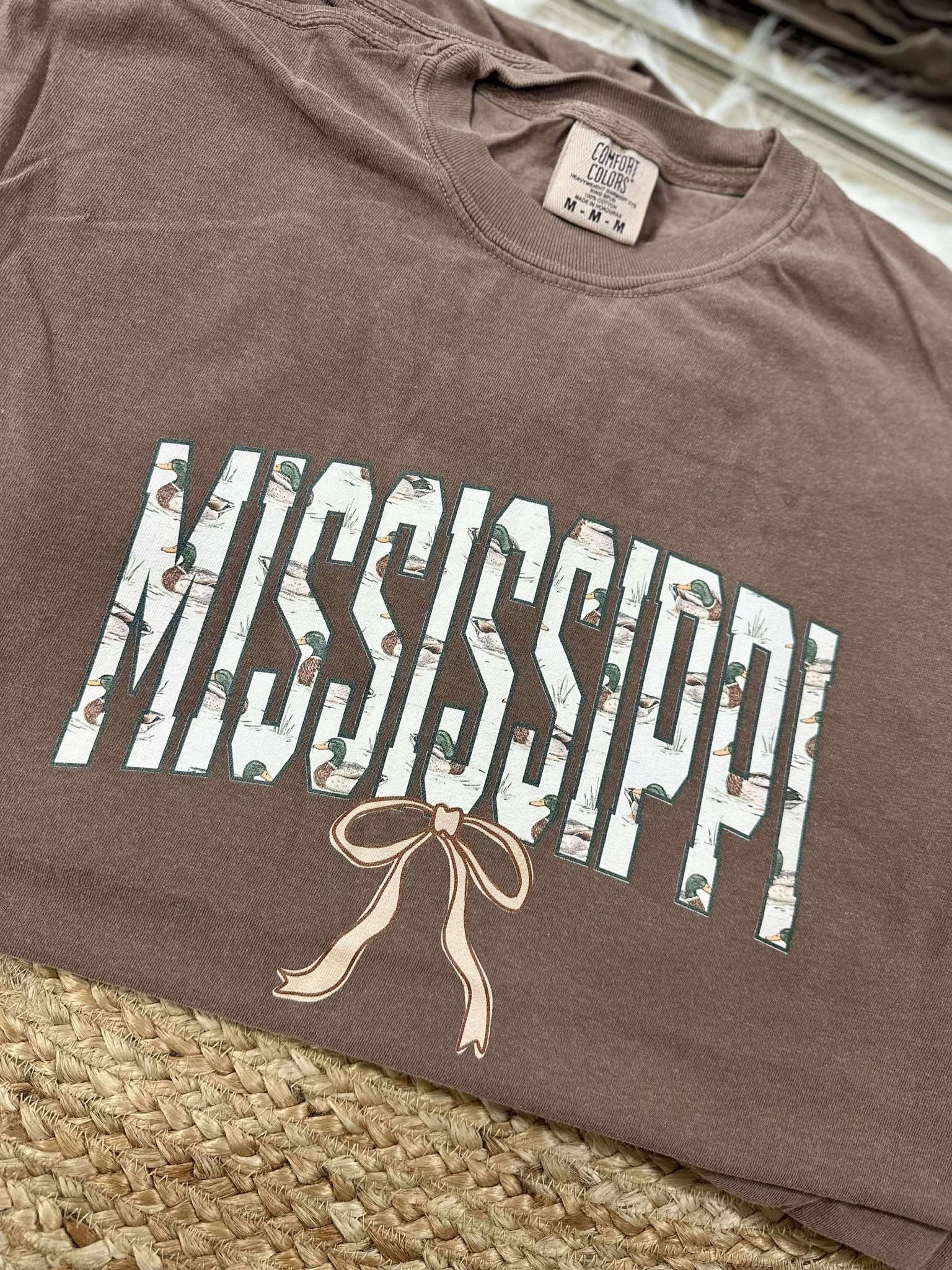 Brown t-shirt with 'Mississippi' printed on it, featuring a bow design, on a woven surface.