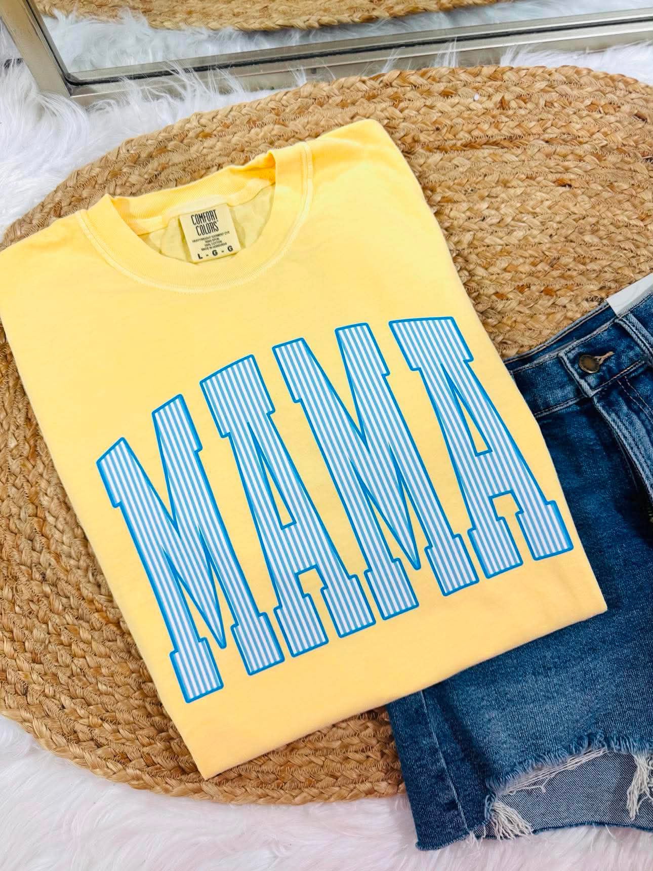 A folded yellow t-shirt with a blue striped 'MAMA' graphic, styled on a woven round mat with blue denim shorts.