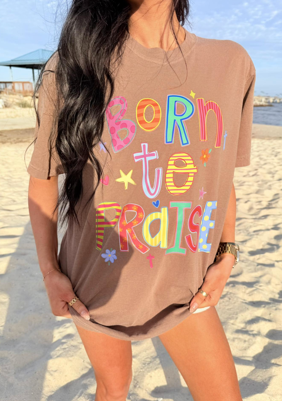 Woman wearing a brown t-shirt with 'Born to Praise' text on a beach