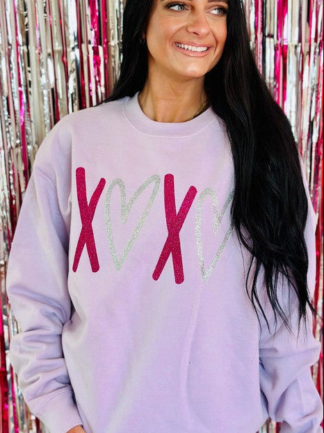 A woman smiling, wearing a lavender multicolor glitter vinyl sweatshirt with a heart and 'XOXO' text on it, standing in front of a pink glittery background.